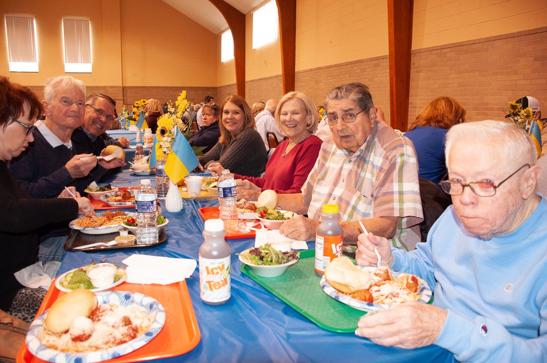 People seated at a long table sharing a meal indoors with blue tablecloths and colorful plates.