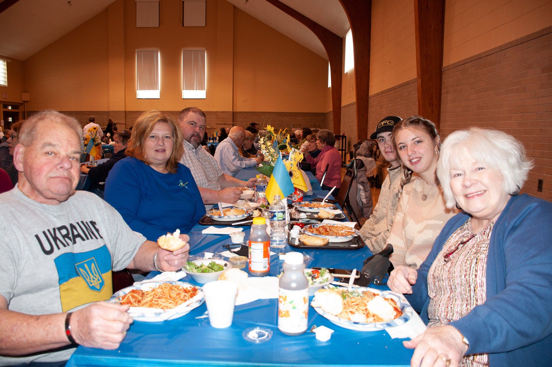 People seated at a long table in a hall, eating a meal and chatting.