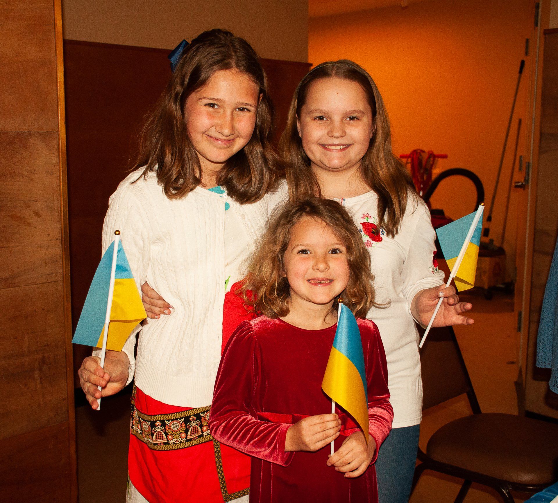 Three smiling children indoors, holding small blue-and-yellow flags.