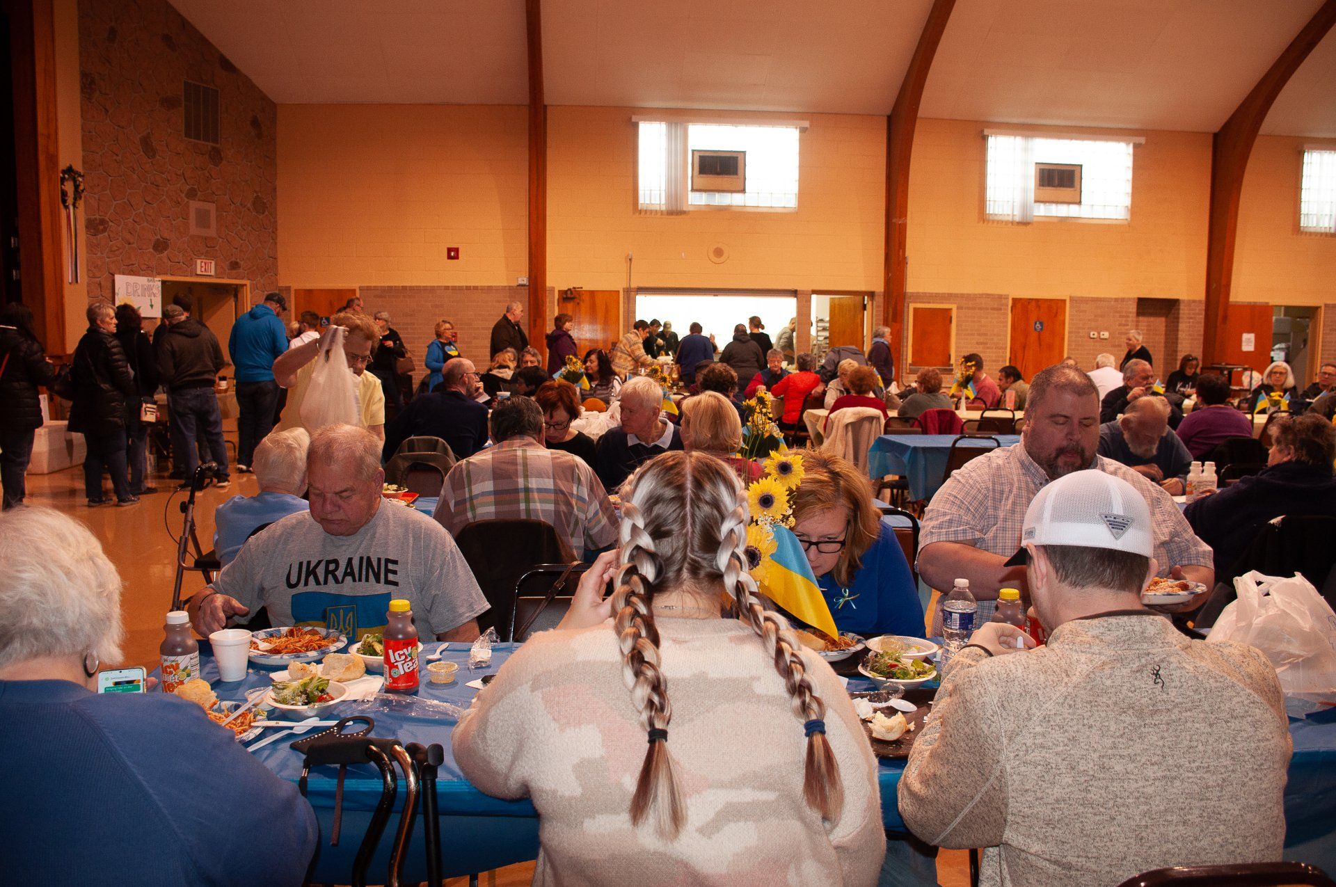 Crowded banquet hall with people seated at long tables, eating and socializing under warm lights