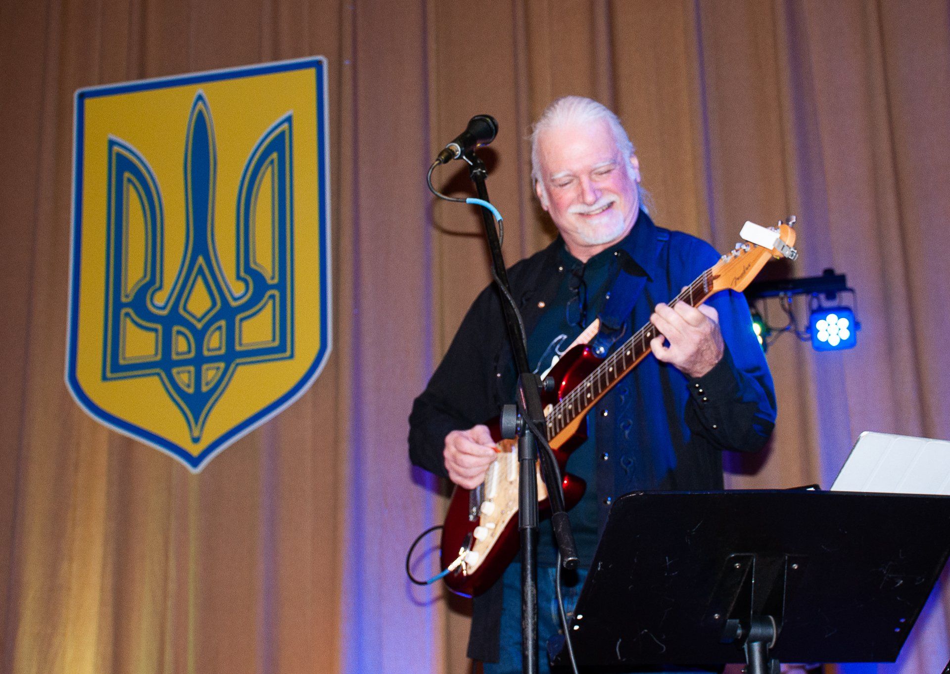 Man playing guitar onstage beside a Ukrainian trident emblem and curtain backdrop