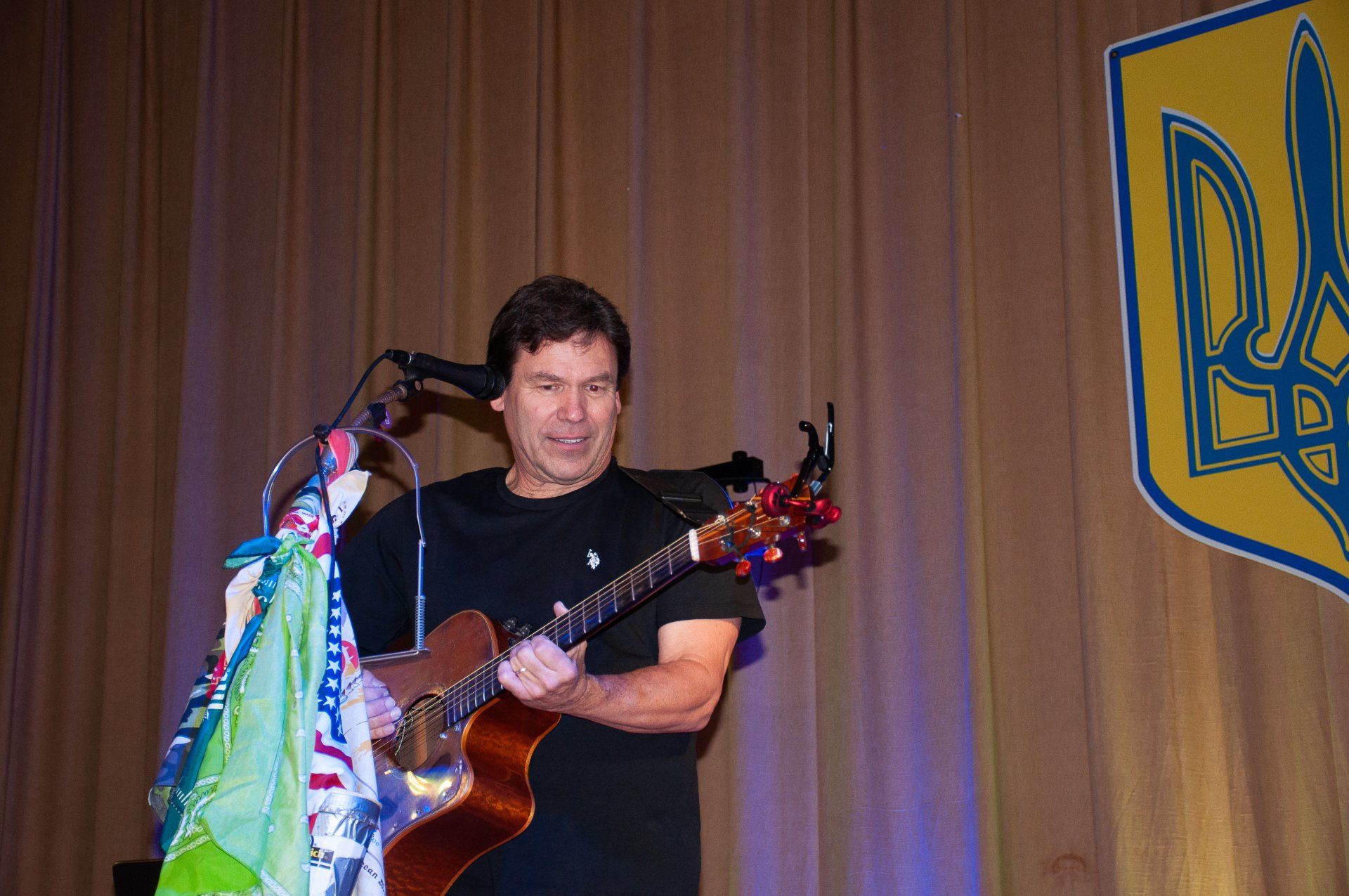Musician singing and playing guitar onstage beside a microphone, with a blue-and-yellow Ukrainian flag backdrop