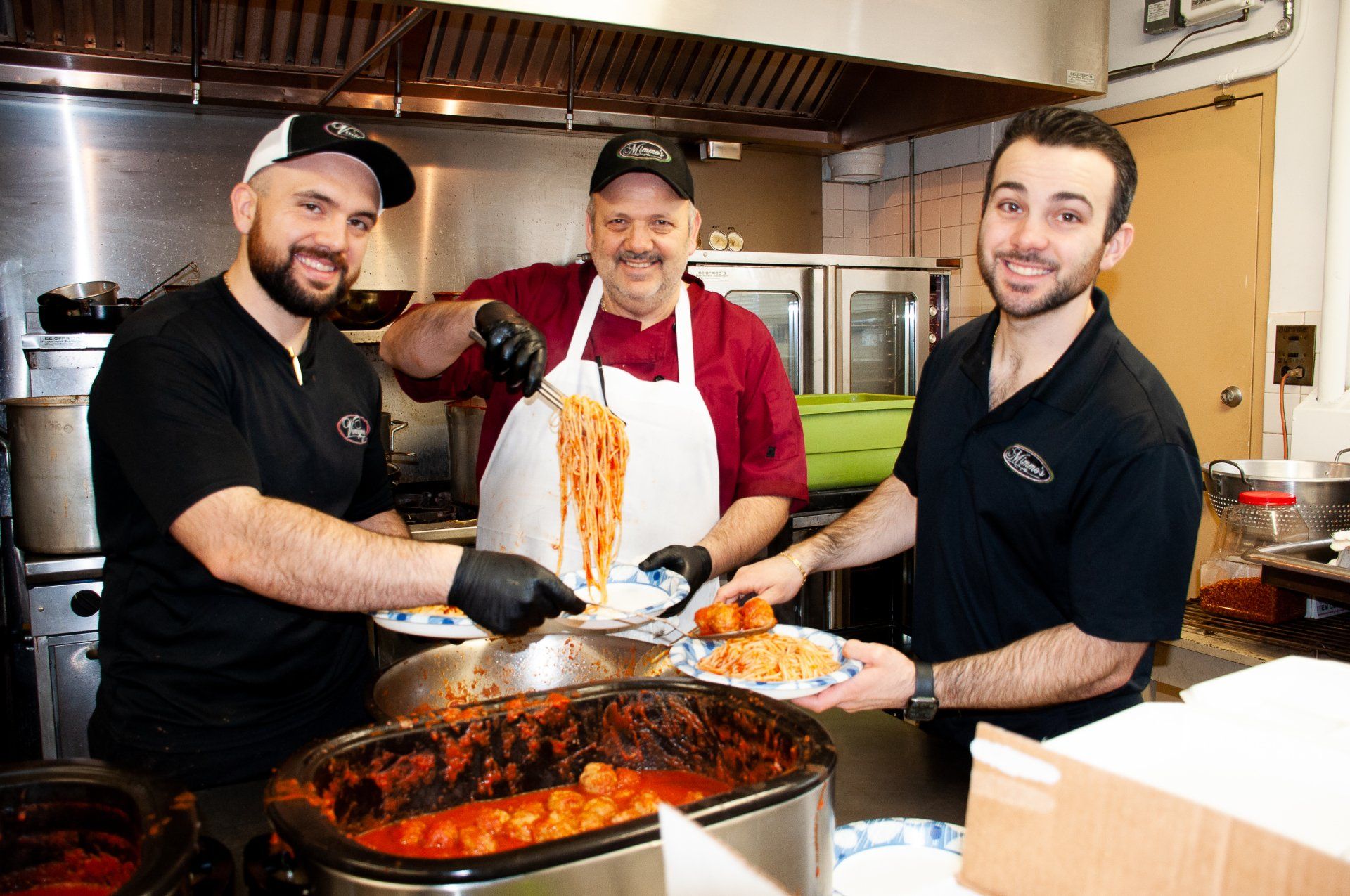 Three chefs in a kitchen serving a pan of pasta and a dish of stew, smiling at the camera