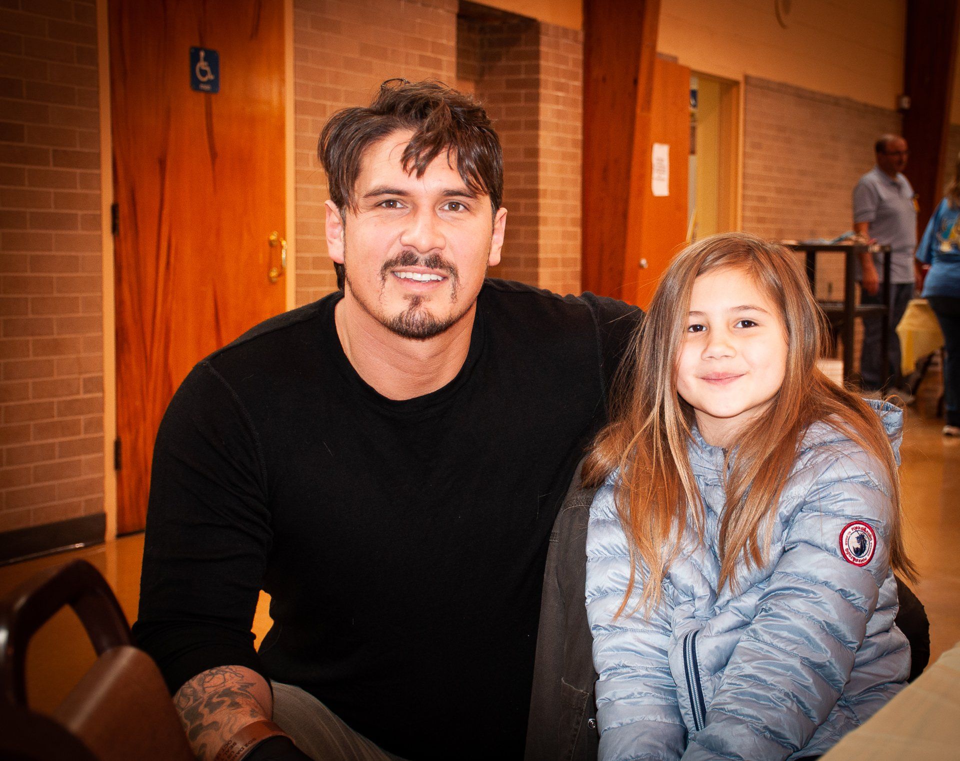 Man and child sitting together in a wood-paneled hallway, both facing the camera and smiling.