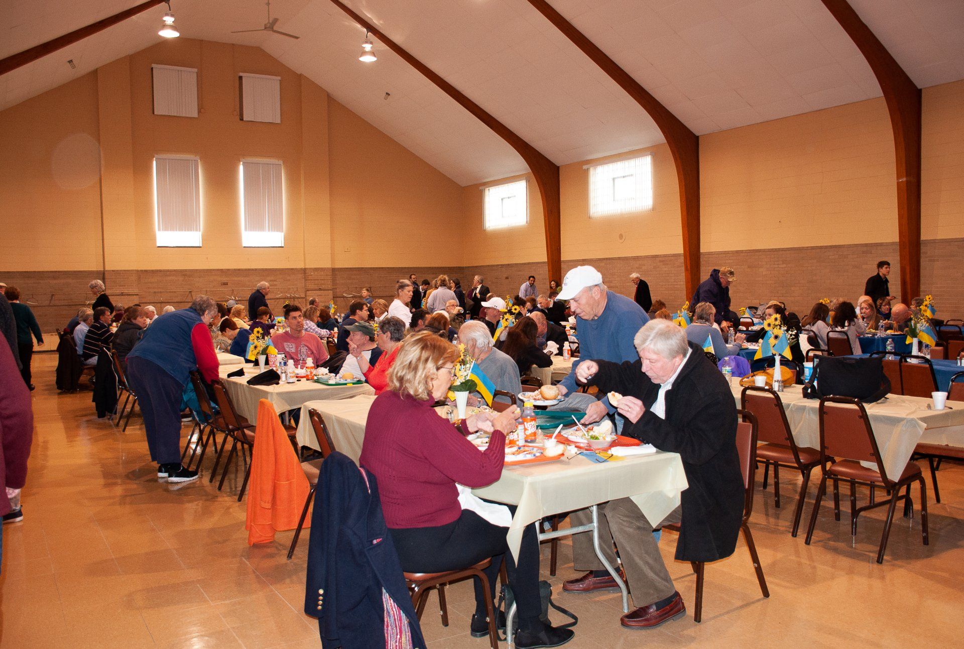 People seated at long tables in a banquet hall, eating and socializing during a community meal
