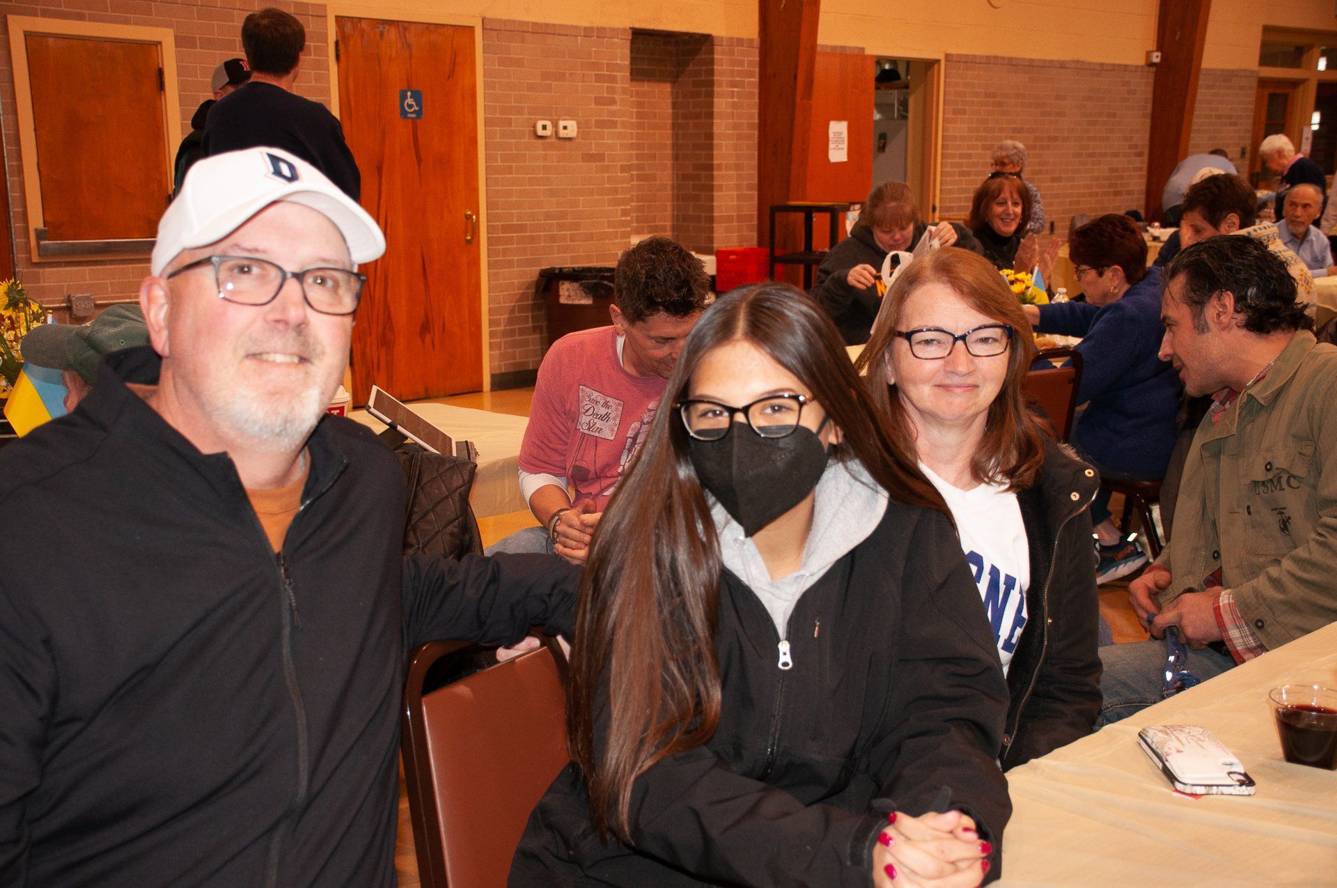 Three people posing indoors at a gathering, one in a black mask and glasses, with tables and seated attendees behind them
