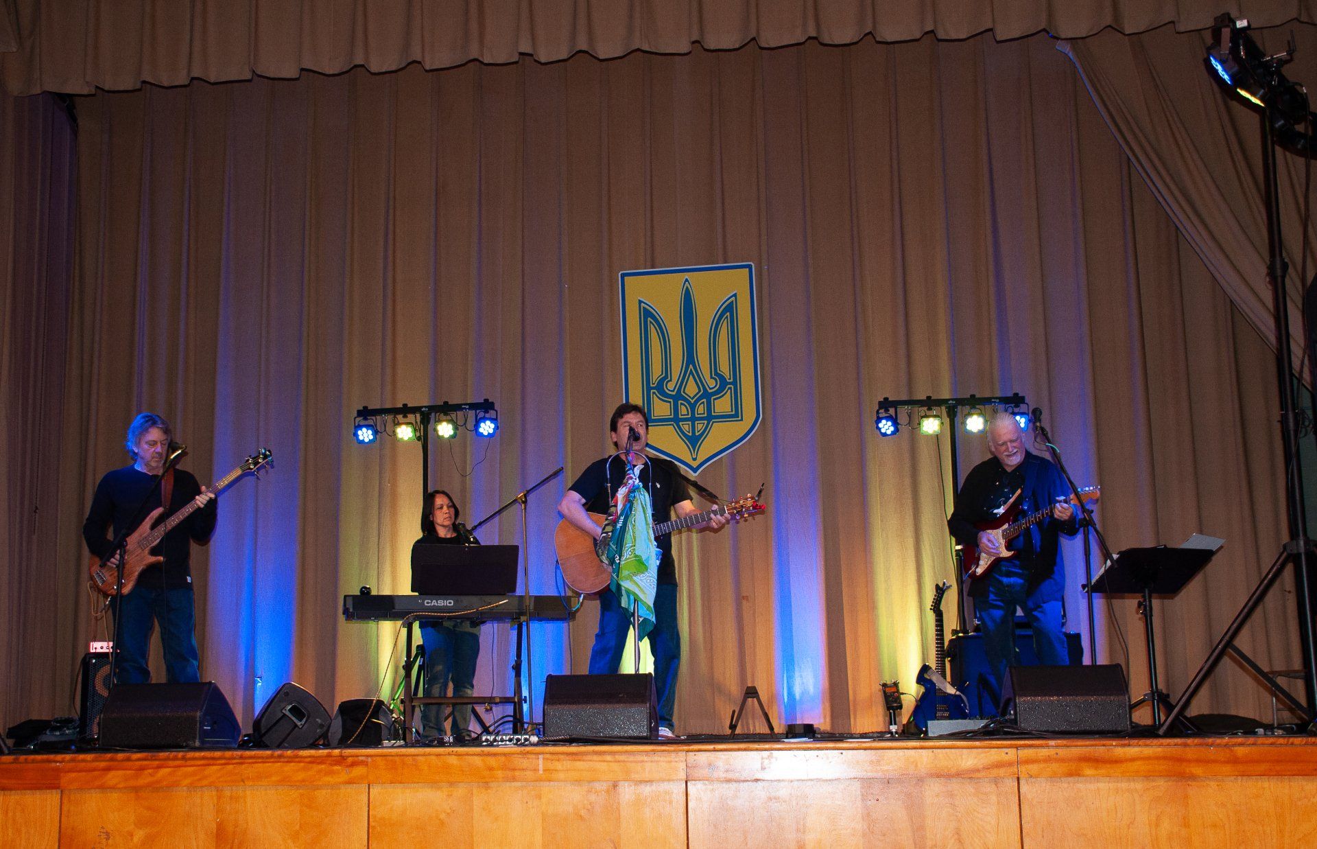 Four-piece band performing onstage under warm lights, with a Ukrainian trident emblem behind them.