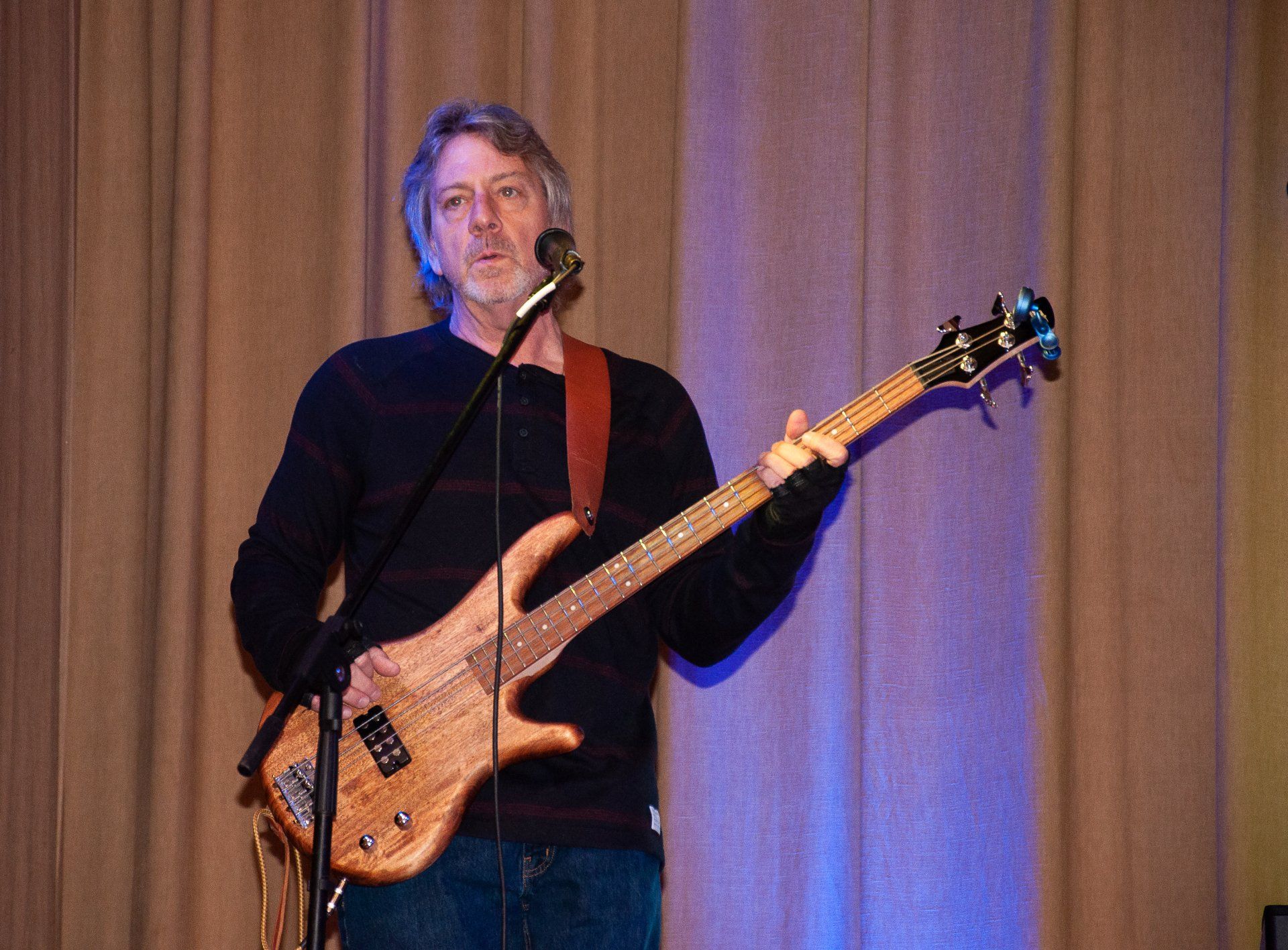 Musician playing bass guitar and singing into a microphone onstage in front of curtains