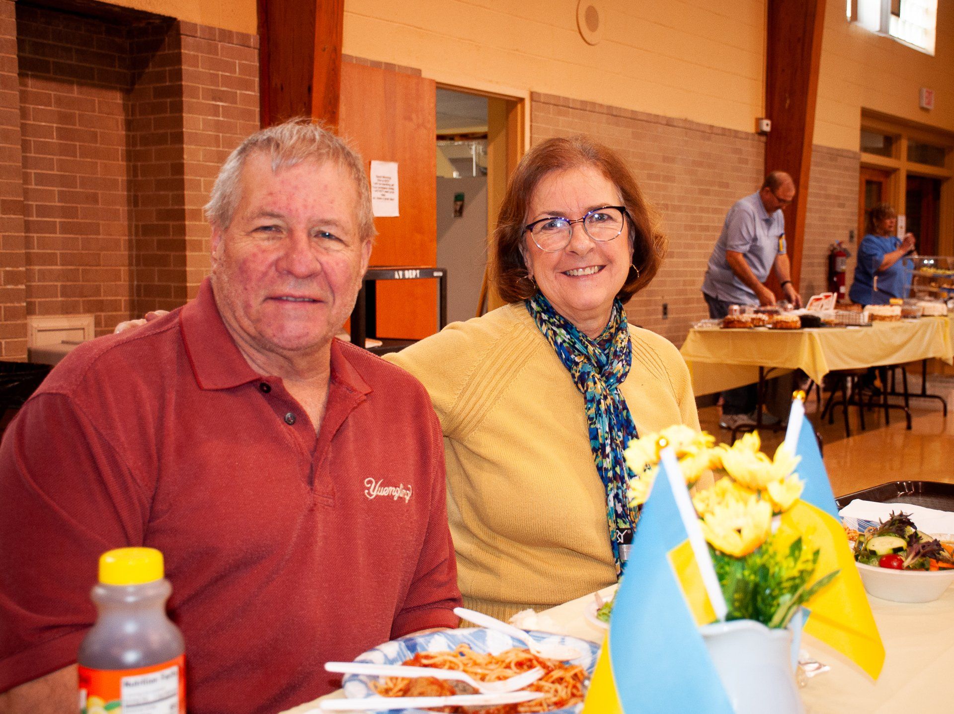 Two people seated at a banquet table with flowers and plates of food in a hall