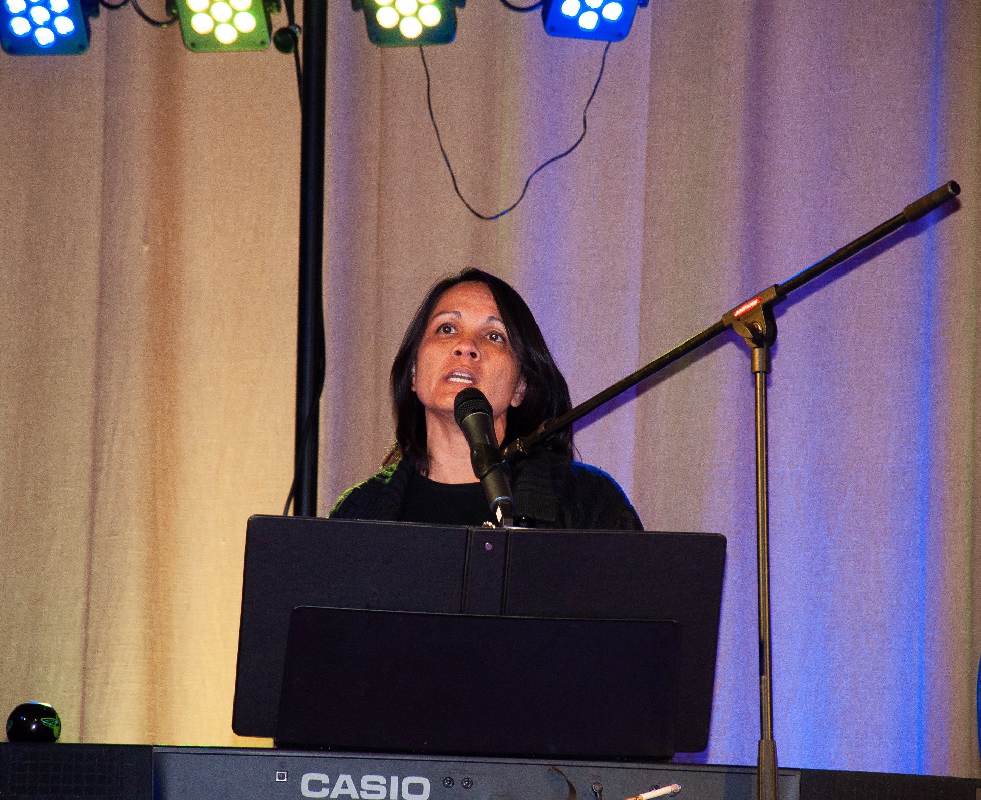 Woman singing into a microphone behind a keyboard on a lit stage