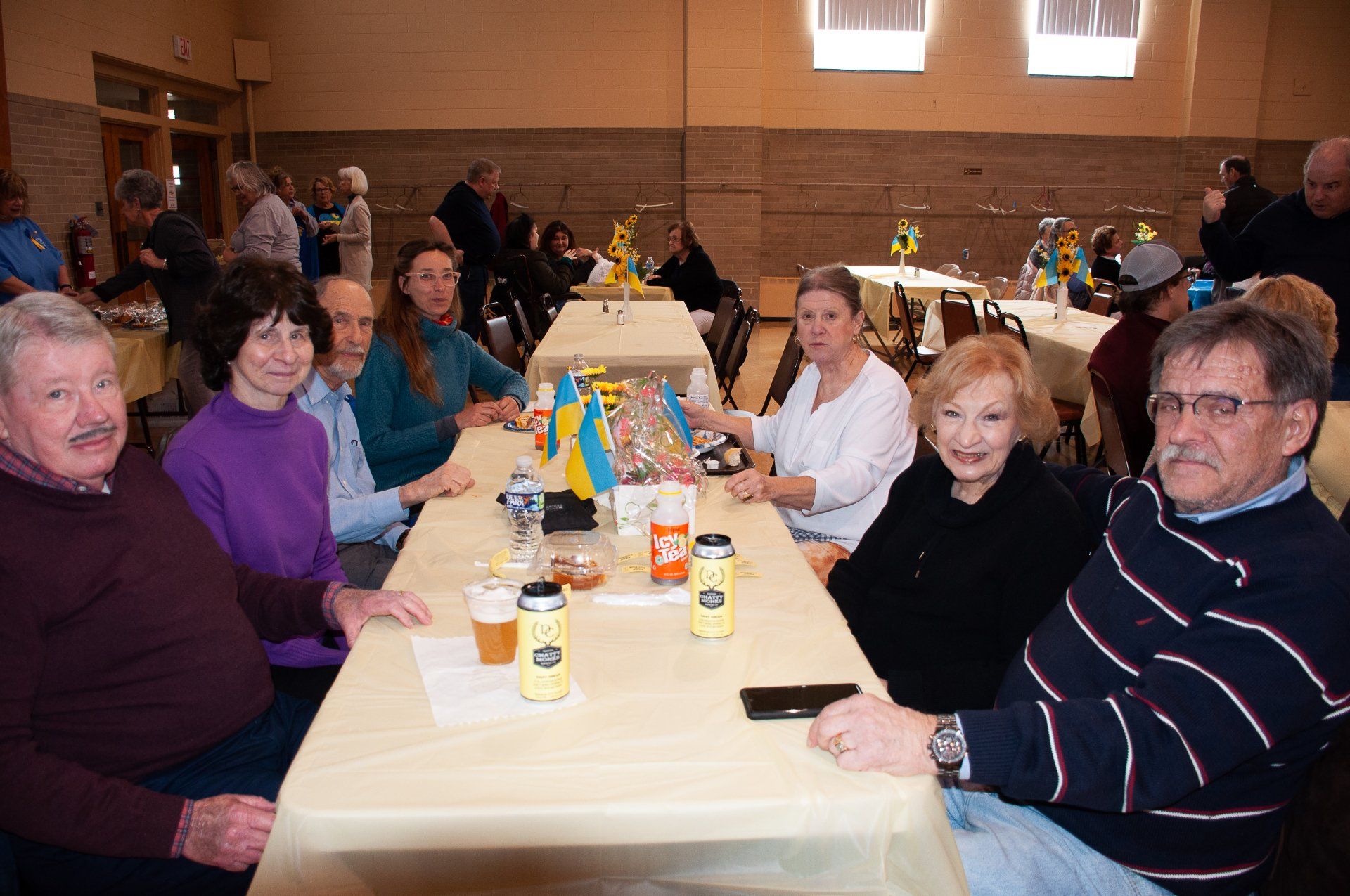 People seated at a long banquet table in a hall, sharing a meal and drinks.