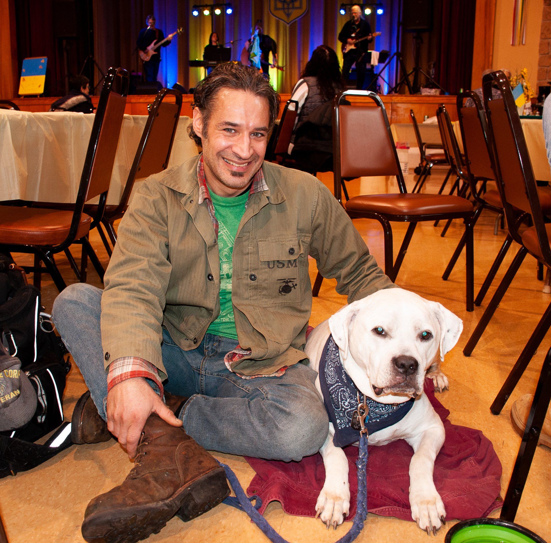 Man sitting cross-legged on a carpet with a white dog in a banquet hall, chairs and stage lights behind them