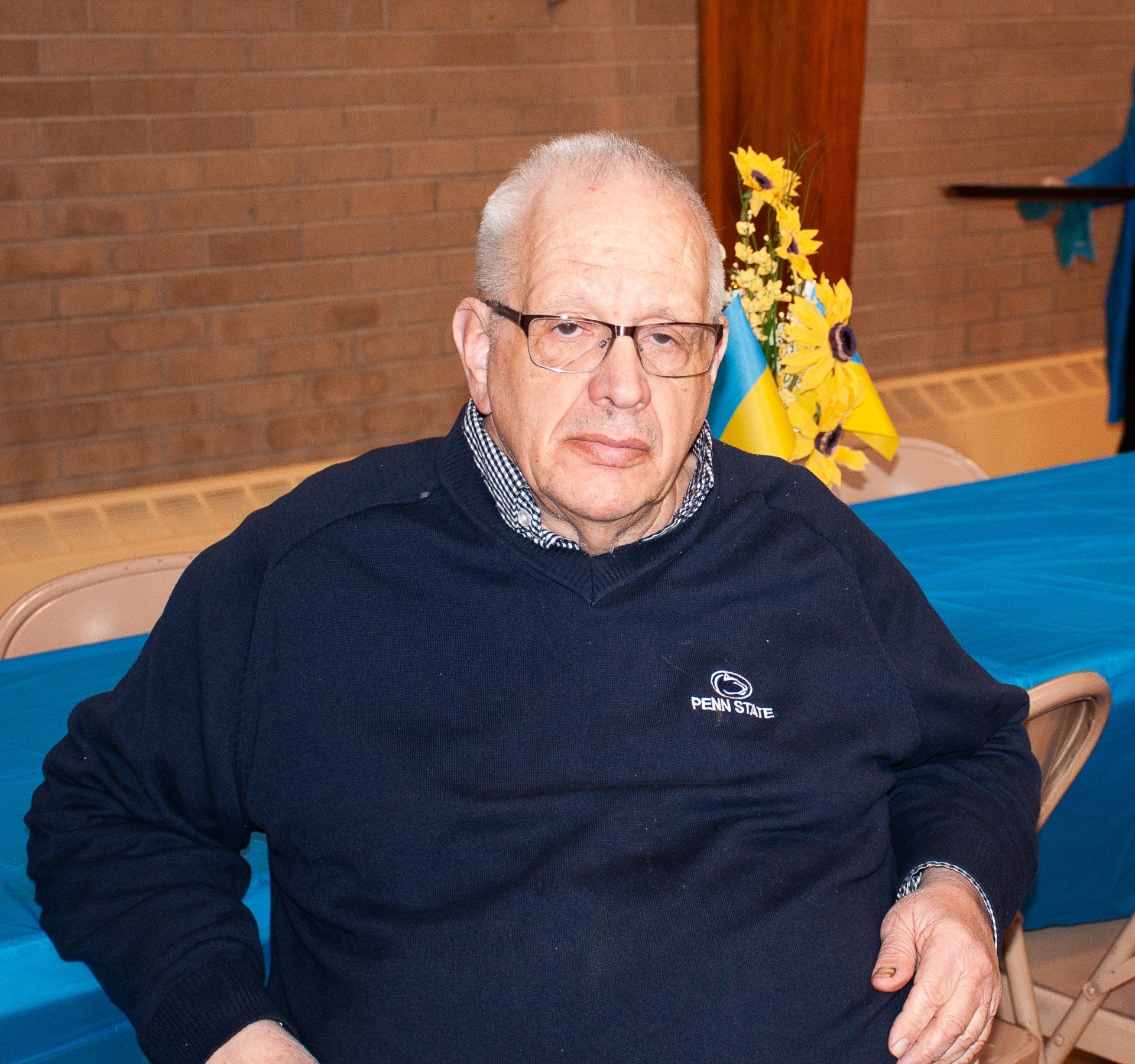 Man seated indoors, wearing glasses and a navy sweater, with a brick wall and flags in the background