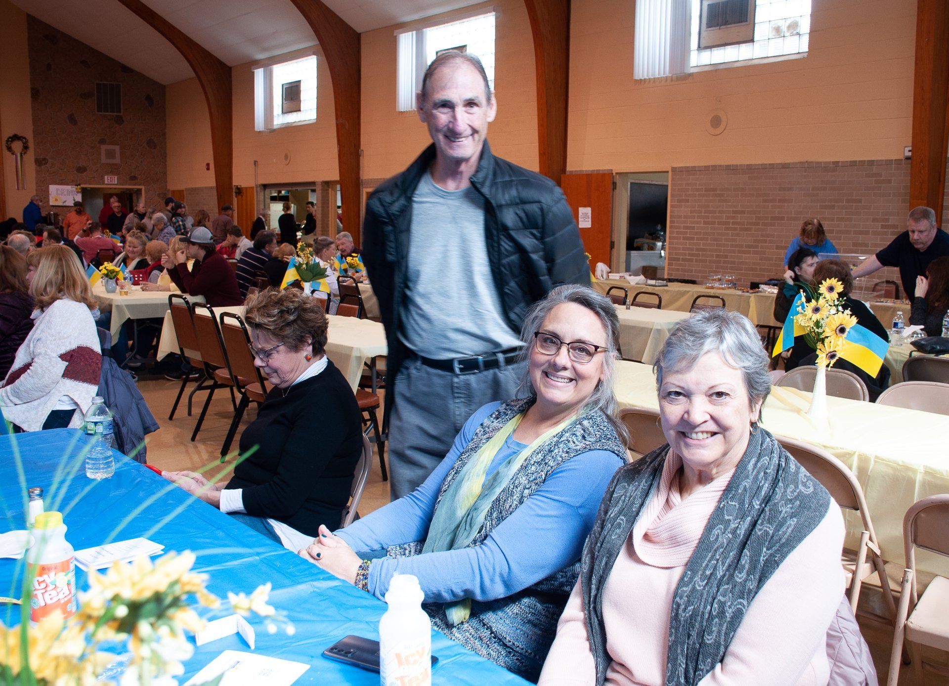 People seated at tables in a community hall, with three smiling guests in the foreground
