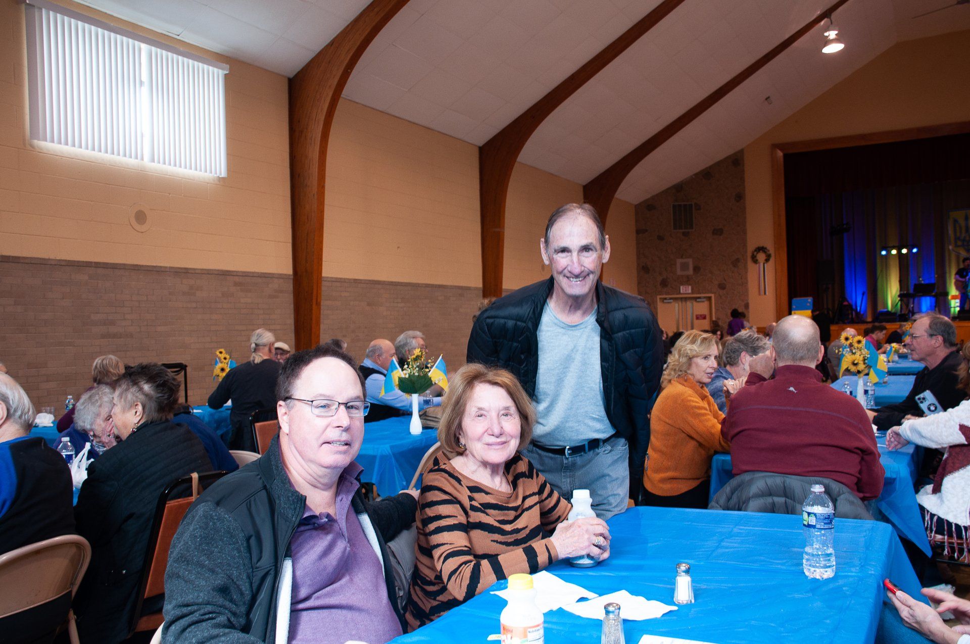 People seated at blue-covered tables in a banquet hall, with a man standing behind them.