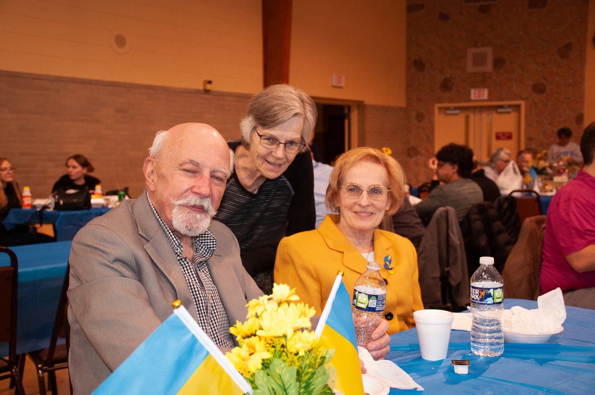 Three people seated at a banquet table with blue tablecloths, smiling with drinks and flowers in front of them.