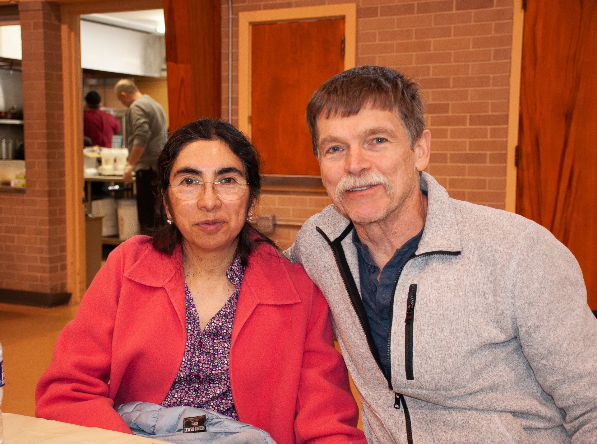 Two people seated indoors, smiling at the camera in a brick-walled room