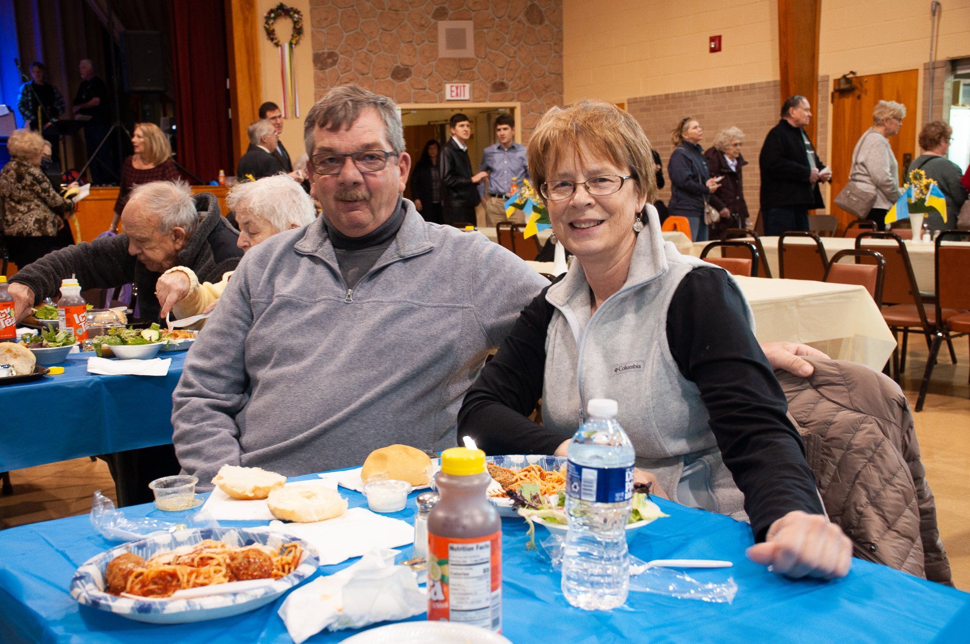 Two people seated at a banquet table with drinks and snacks in a community hall