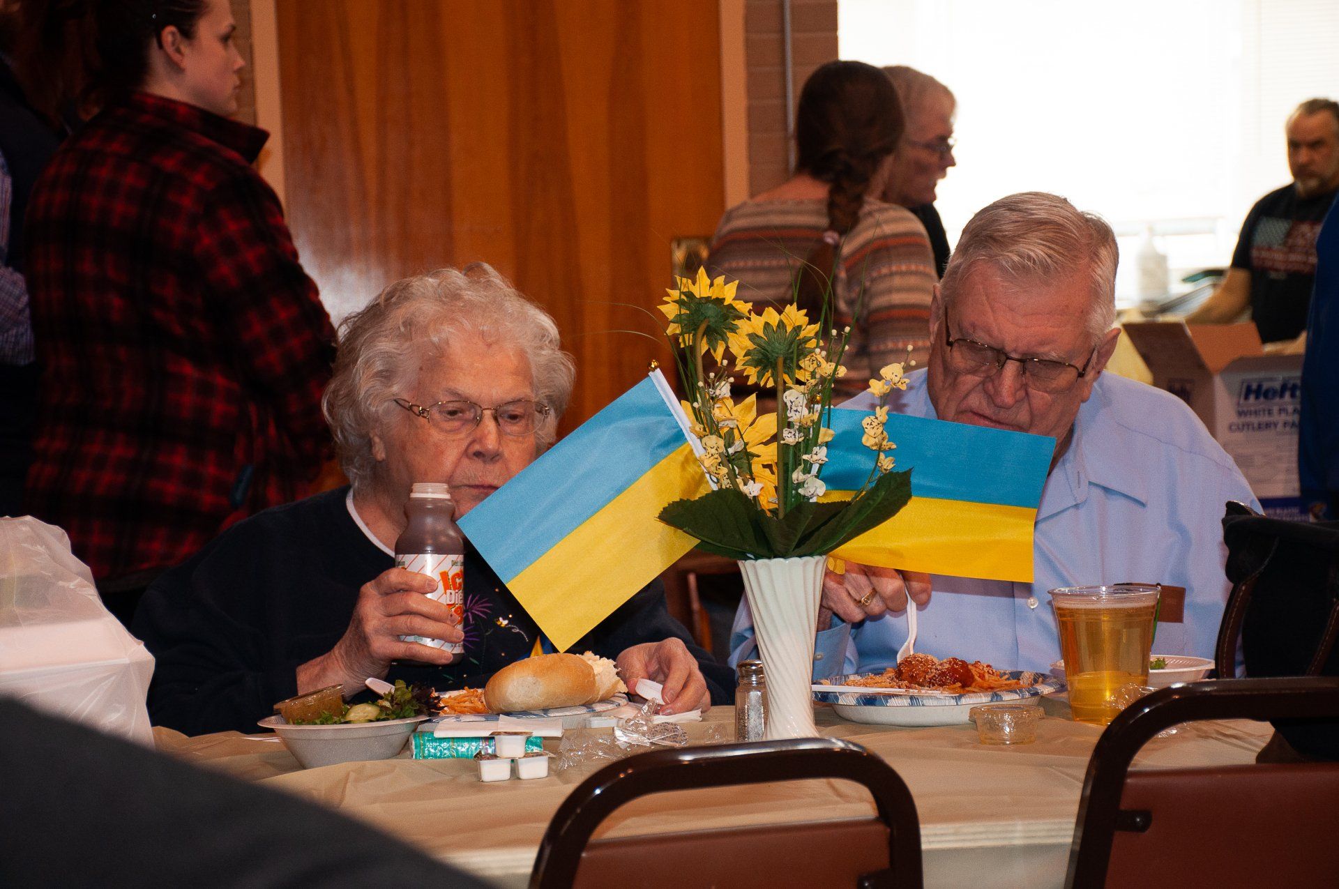 Two elderly people at a table with blue and yellow flags and flowers in a community hall