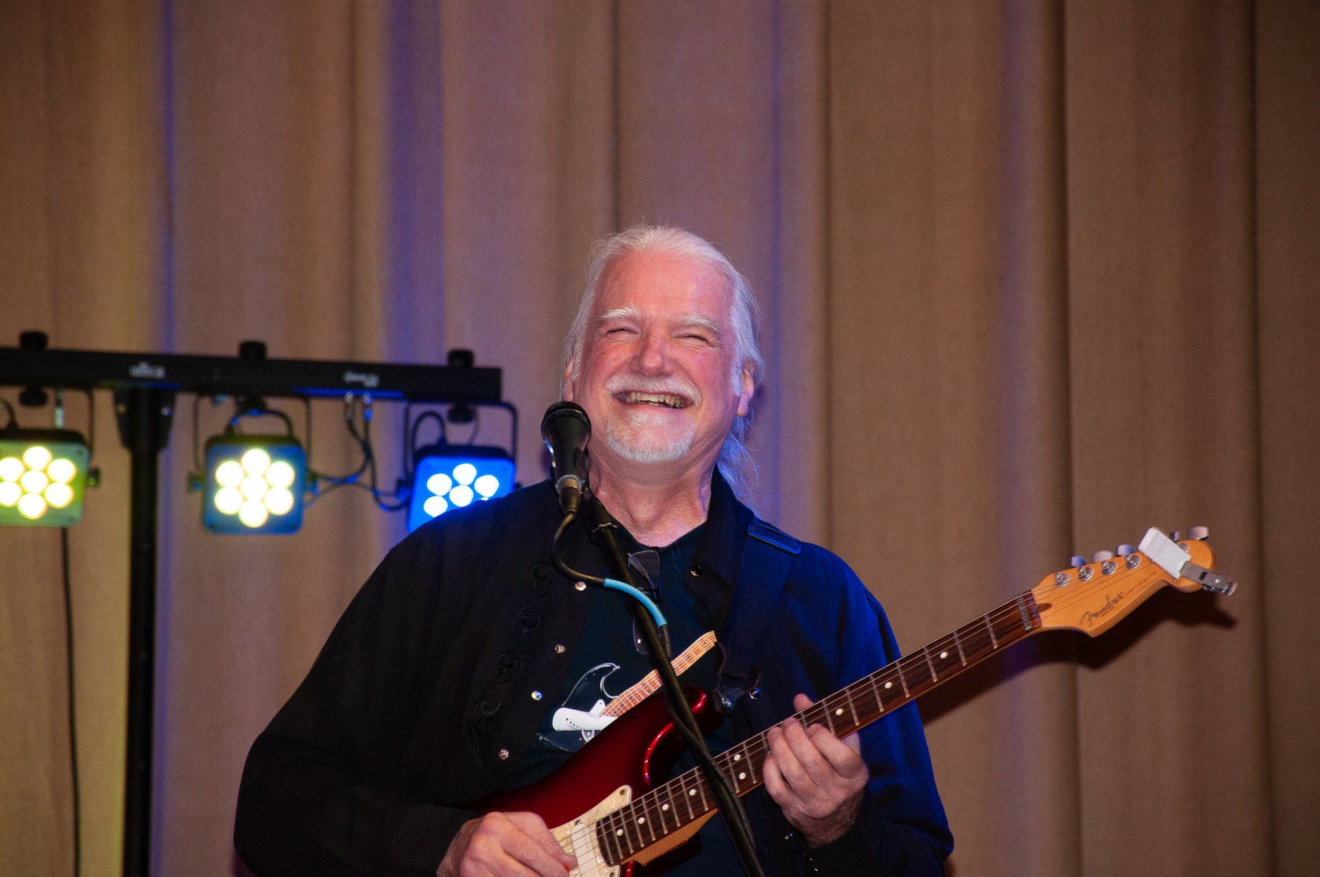 Smiling guitarist on stage under blue lights, playing an electric guitar in front of curtains