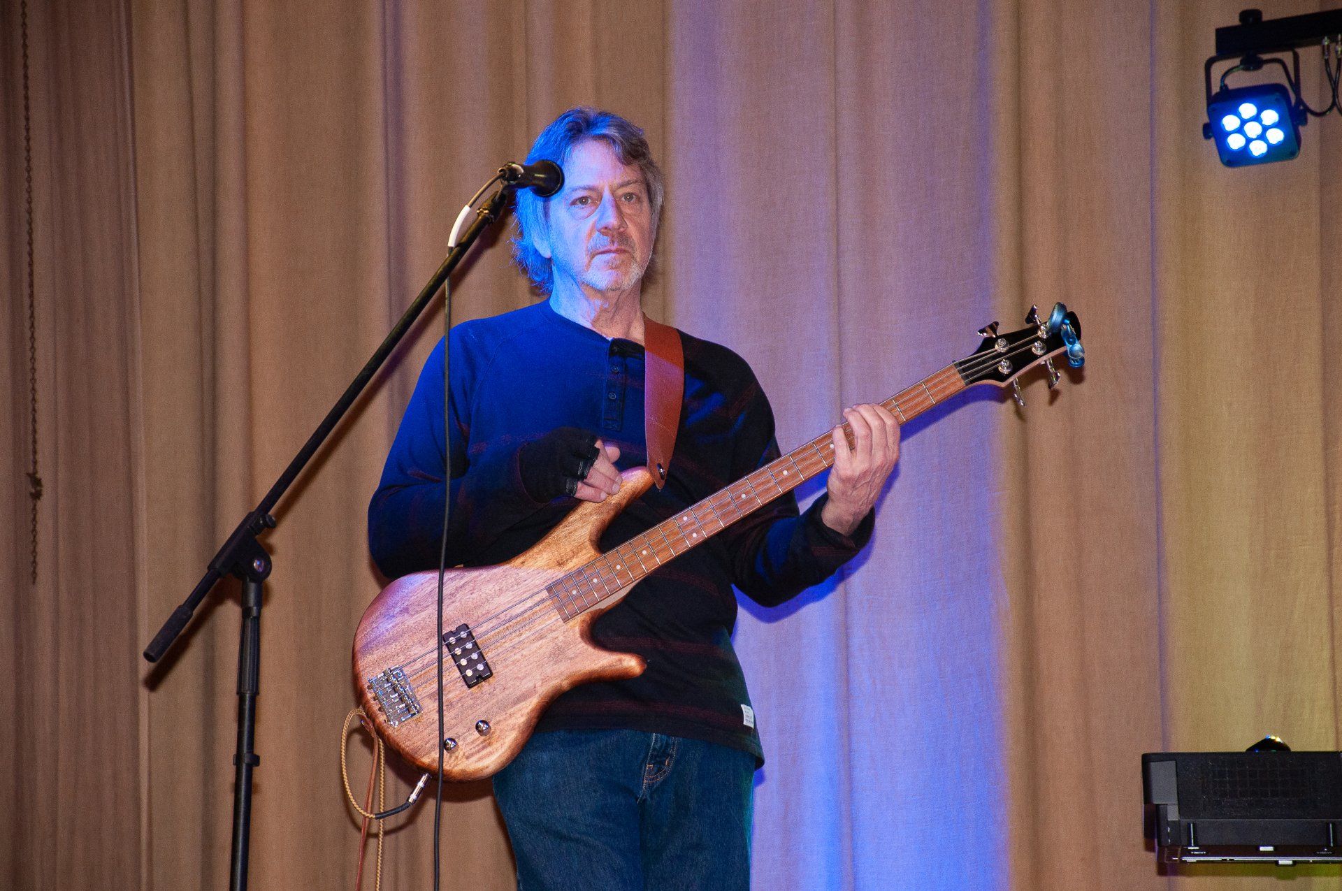 Musician playing bass guitar onstage under blue and purple lights, standing beside a microphone stand.