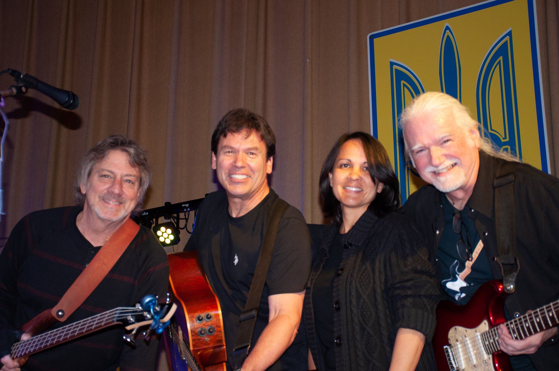 Four musicians posing with guitars onstage in front of a blue-and-yellow banner