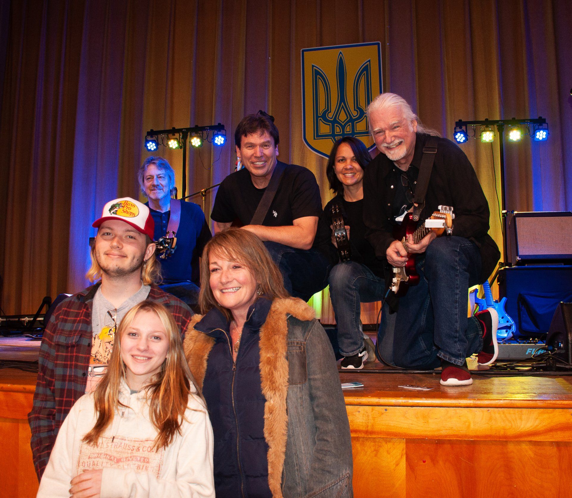 Group posing on a stage with a blue-and-gold backdrop, smiling and kneeling in front of instruments.