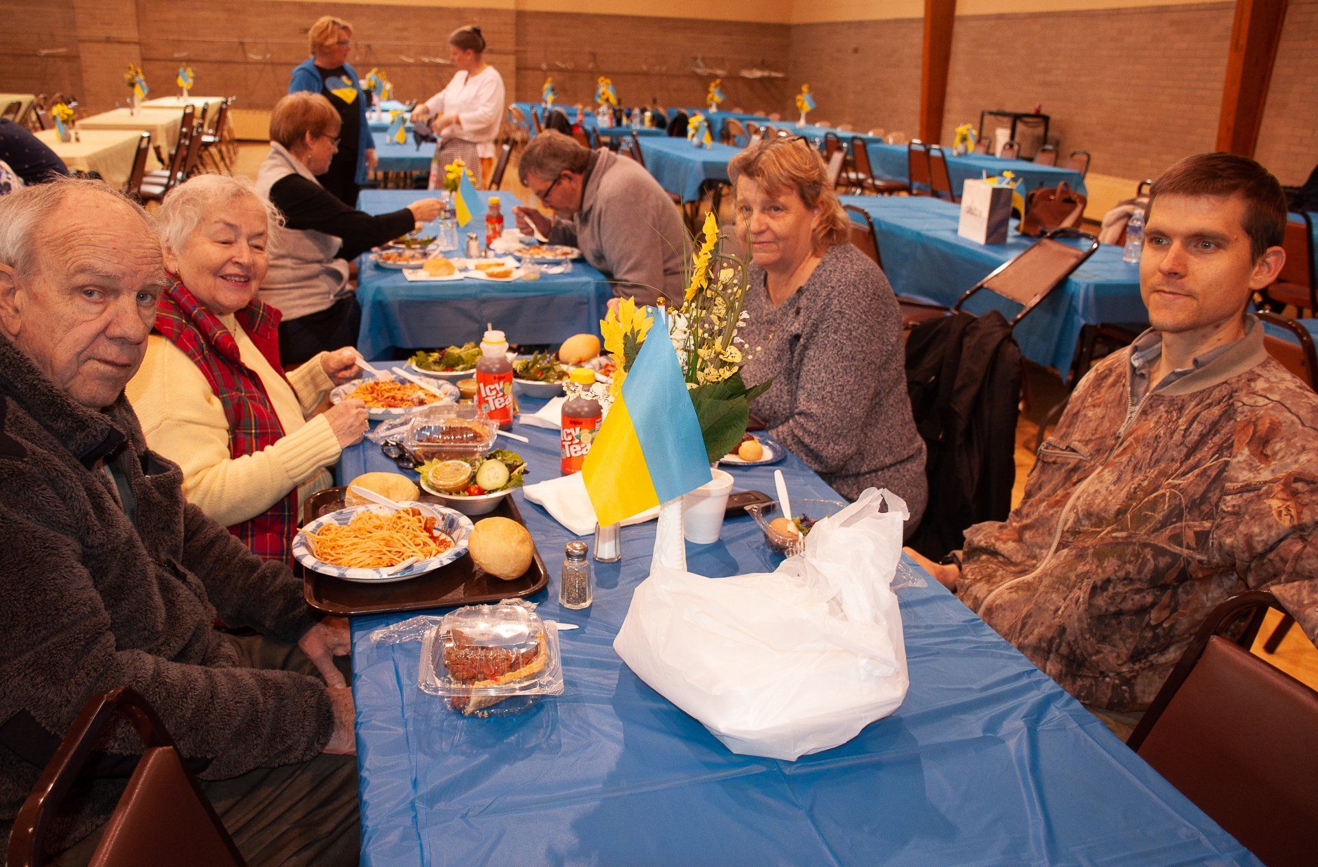 People seated at a banquet table with blue tablecloths, food, and a small Ukrainian flag in a hall