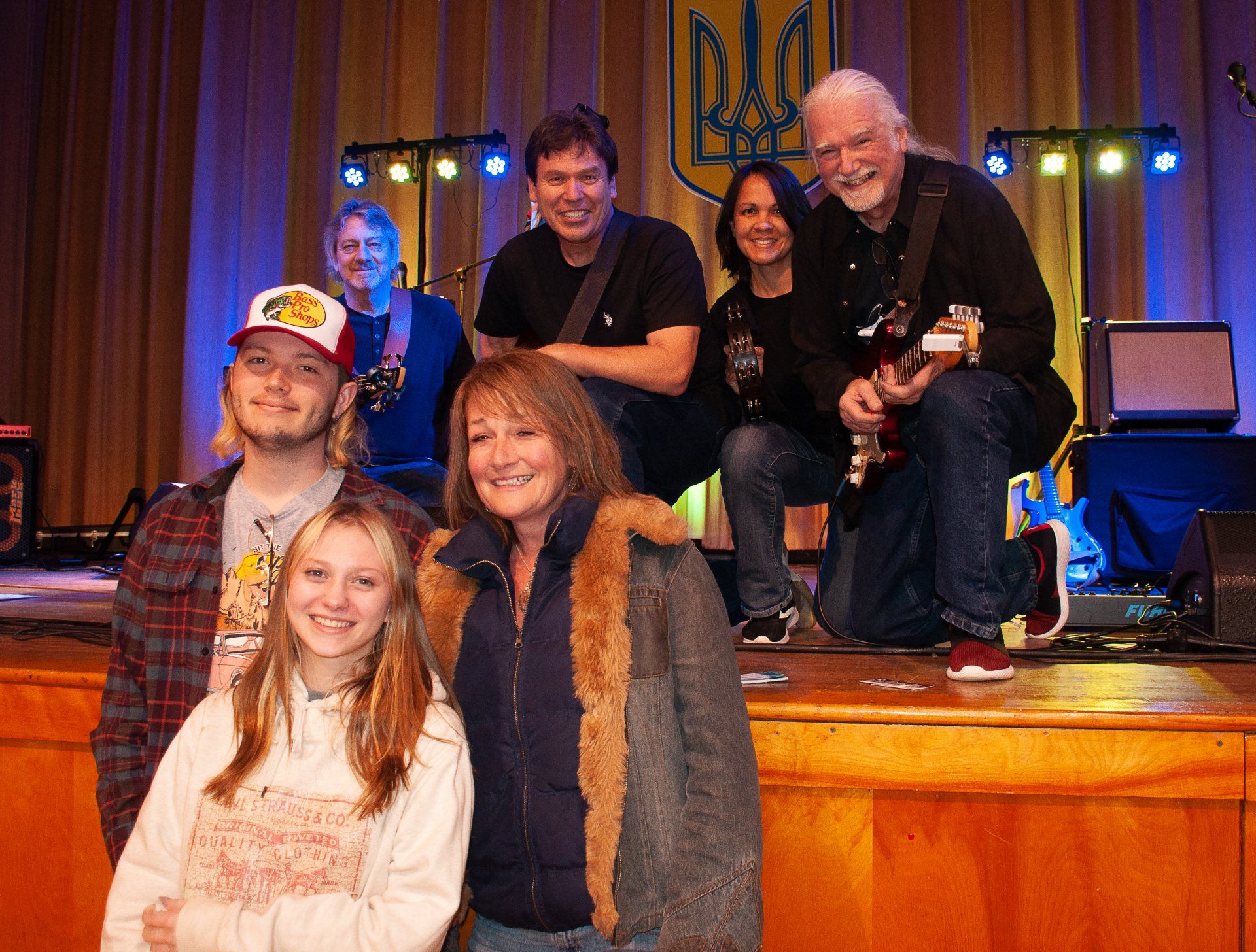 Group of smiling people posing on a stage with blue curtains and warm lighting, one holding a violin.