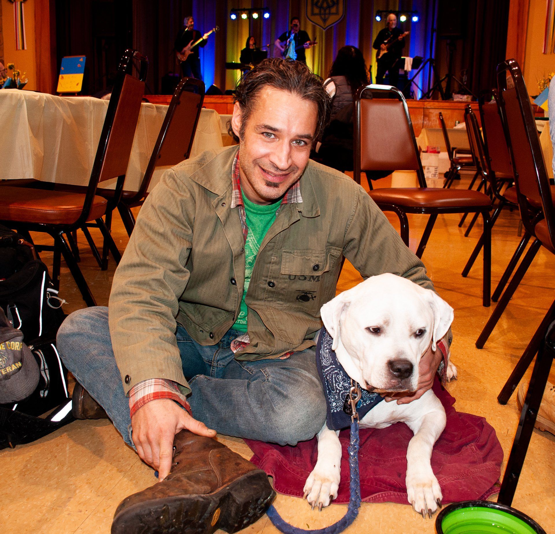 Man sitting on the floor with a white dog on a blanket in a warmly lit room with chairs and tables.