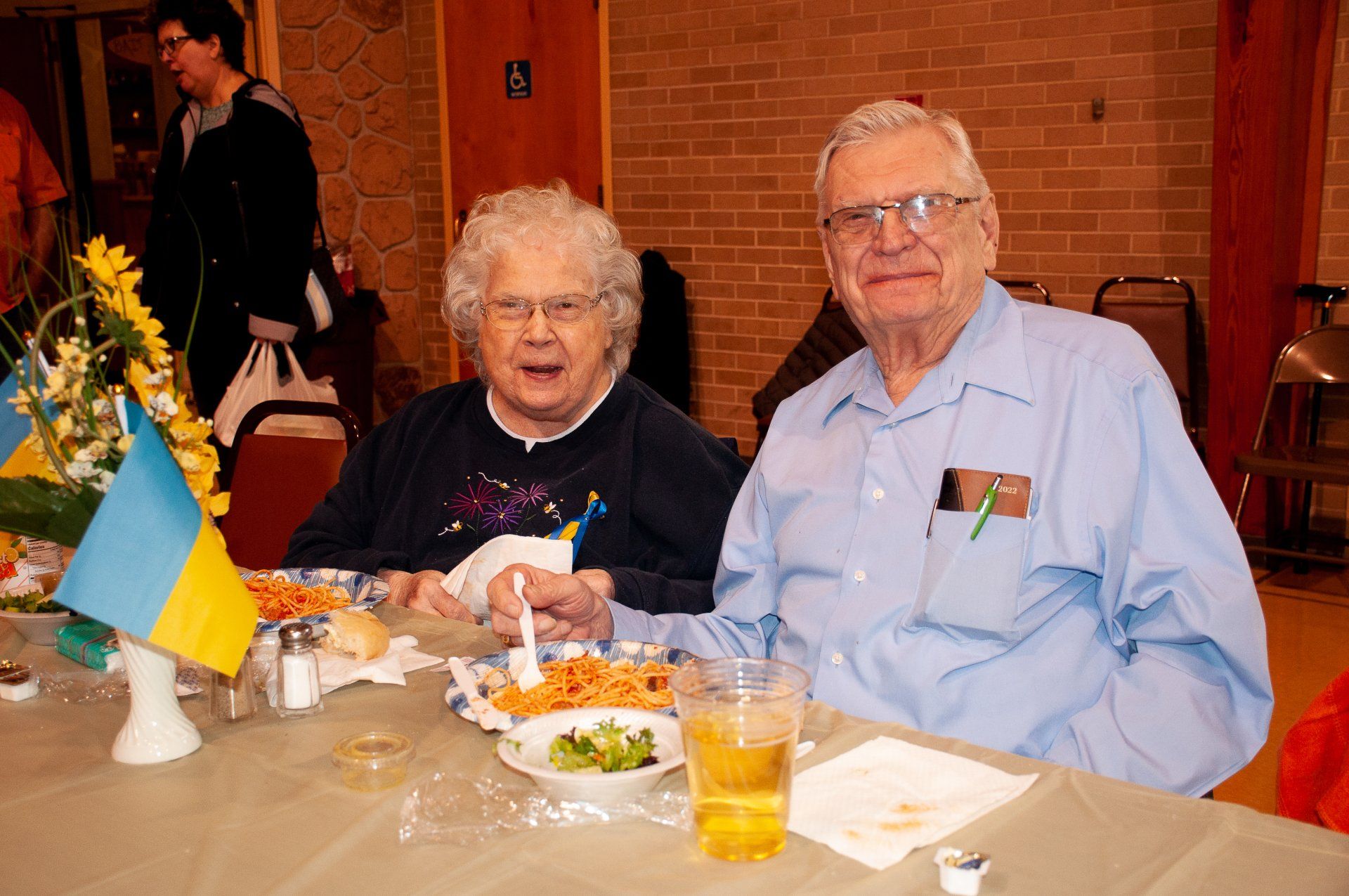 Two people seated at a dining table with drinks, food, and a small blue-yellow flag in a warm restaurant setting