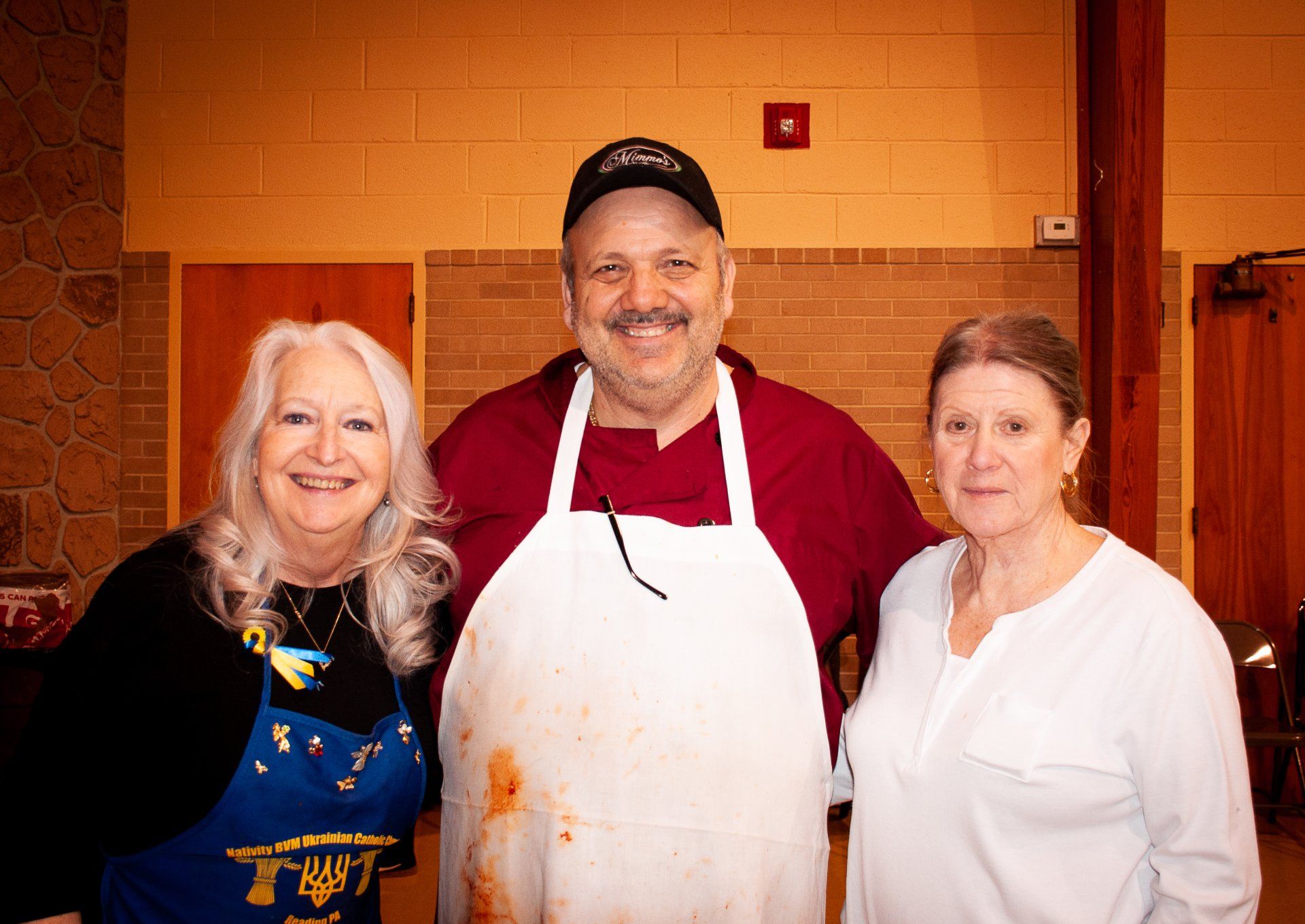 Three smiling people in aprons stand together in a kitchen.