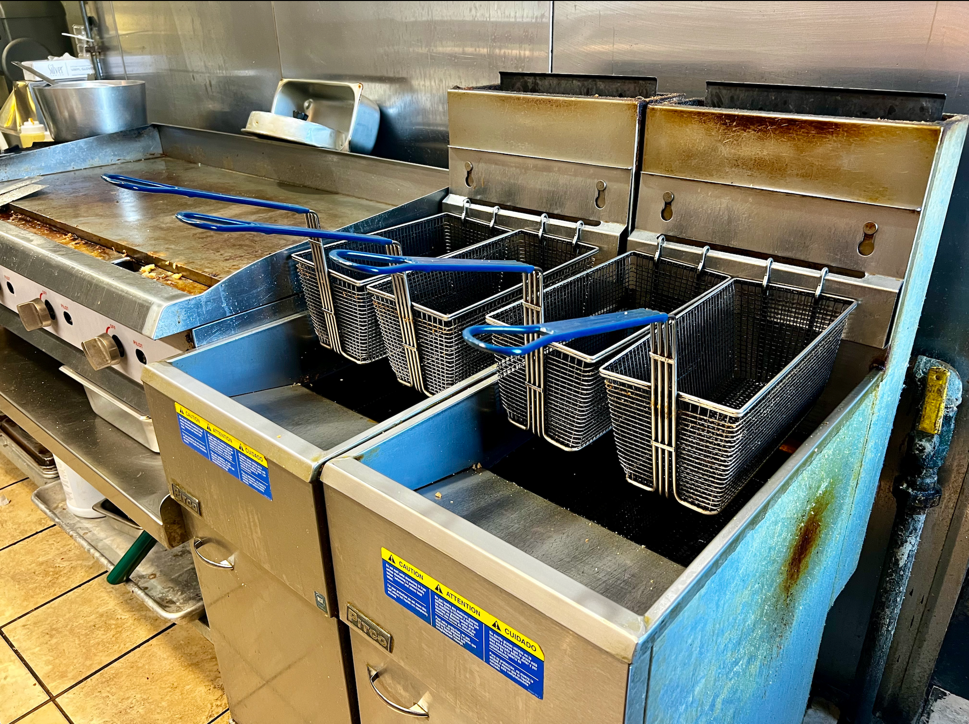 Stainless steel deep fryers with baskets in a commercial kitchen, stained with use.