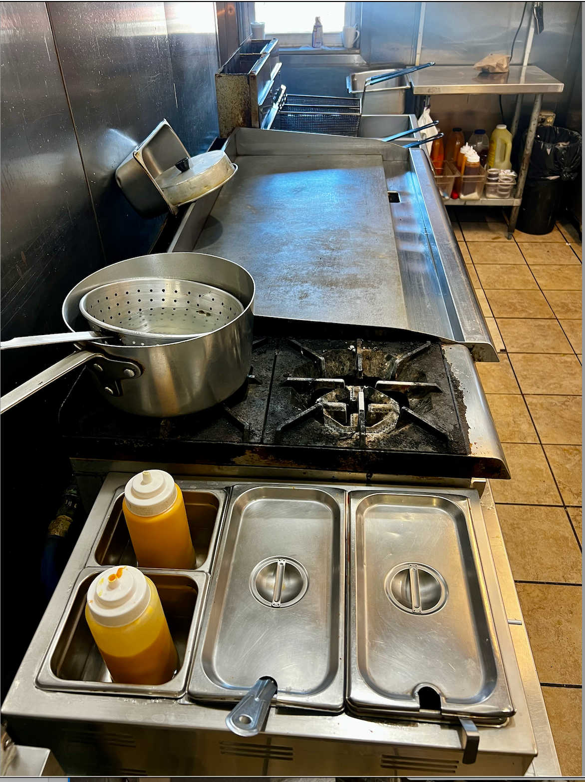 Commercial kitchen stovetop with pot, condiment bottles, and metal prep table under stainless steel counters