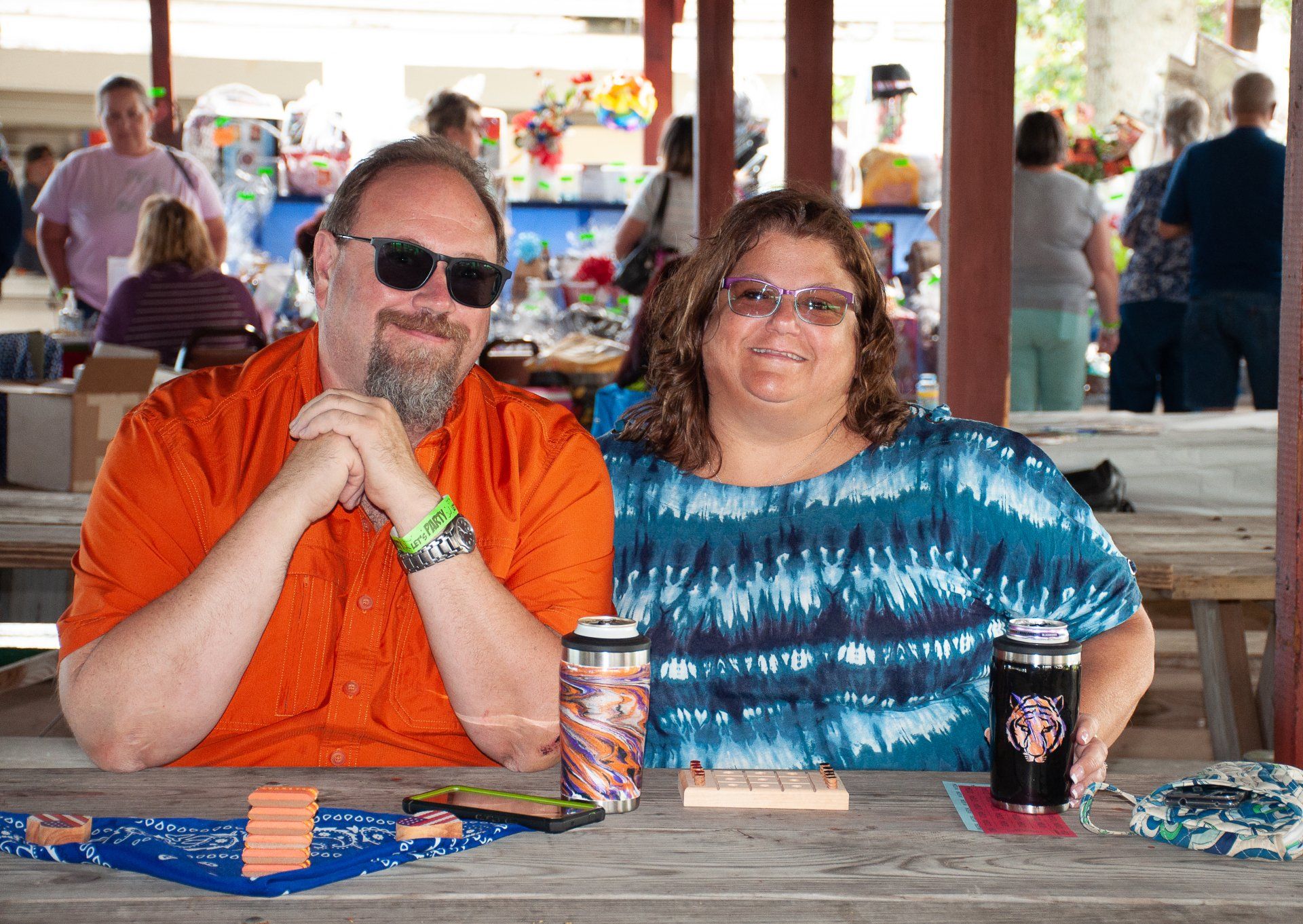 Two people seated at a picnic table, smiling in a covered outdoor pavilion with drinks and snacks