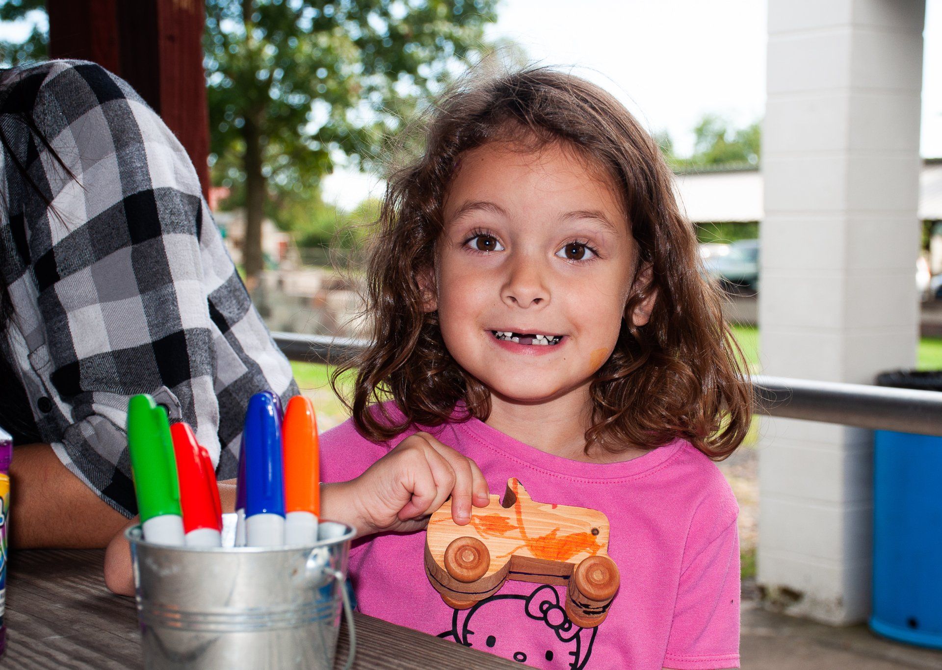 Smiling child in pink shirt holding an orange toy near a cup of colorful markers outdoors
