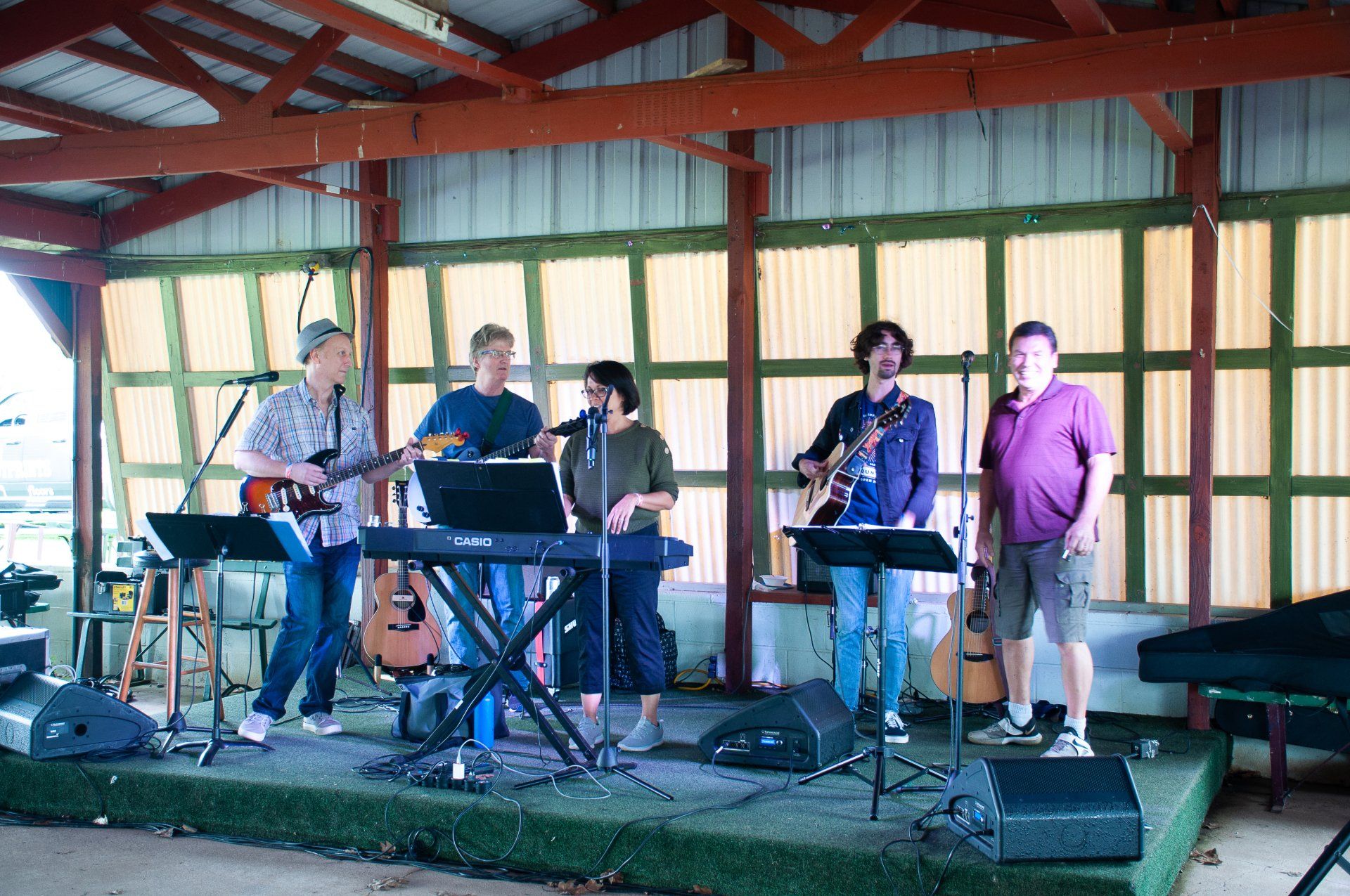 Five people on an outdoor stage, some playing instruments, under a red wooden pavilion