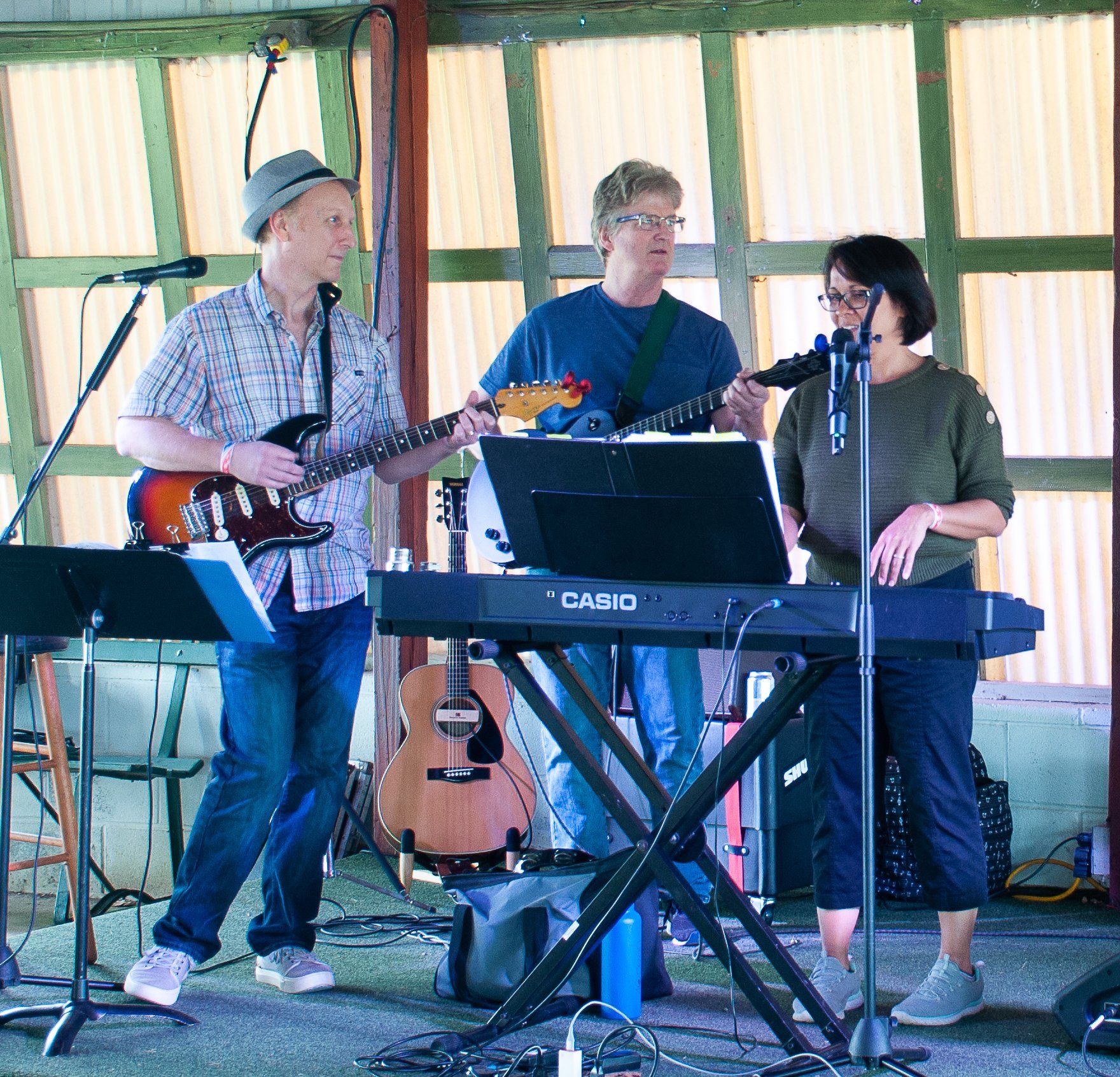 Three musicians performing indoors, with guitar, keyboard, and microphones on a small stage.