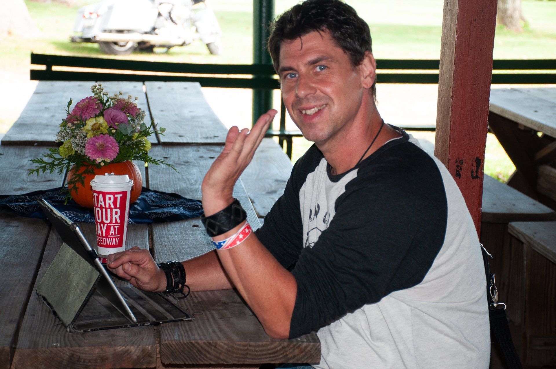 Man smiling and waving at a table with a laptop, next to a red book in a cafe