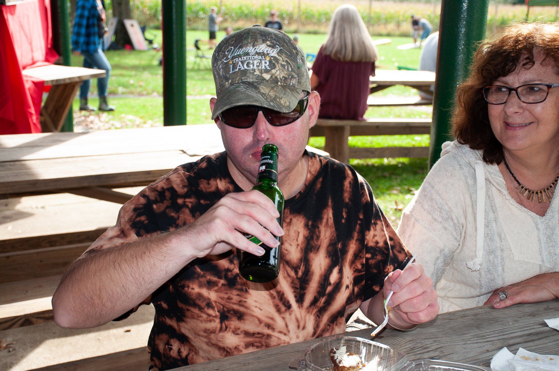 Man in camo cap drinks from a bottle at a picnic table beside a woman in an outdoor pavilion.