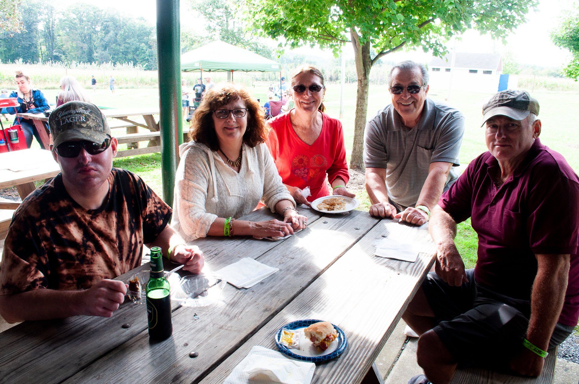 Six people seated at a picnic table outdoors, smiling and talking under a shaded pavilion.