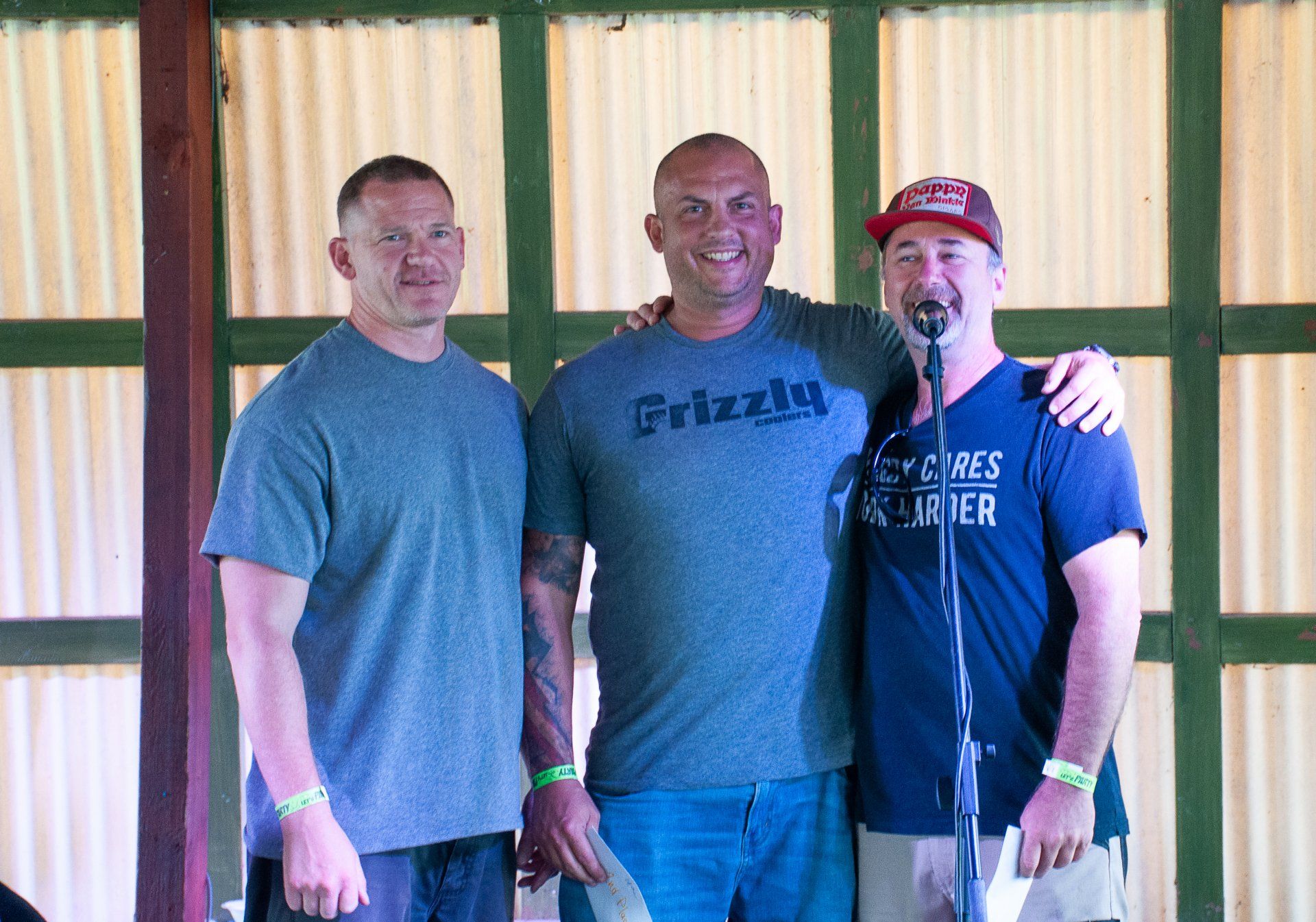 Three men standing together indoors, with one speaking into a microphone in front of a striped wall.