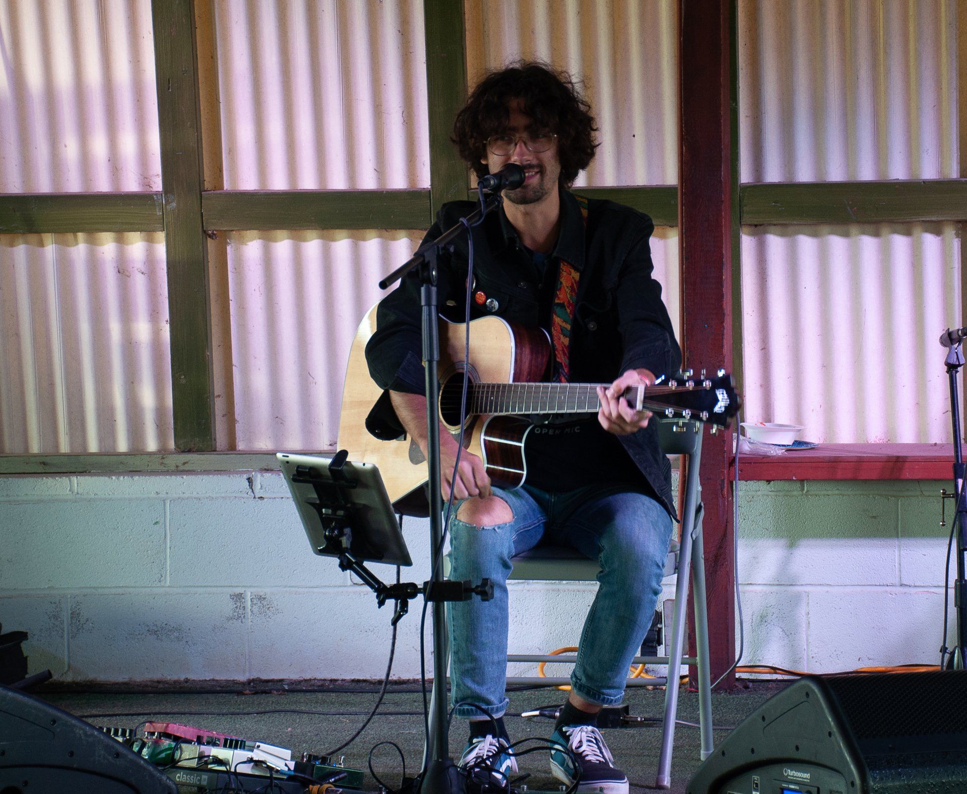 Musician seated on a stage, singing into a microphone while playing acoustic guitar