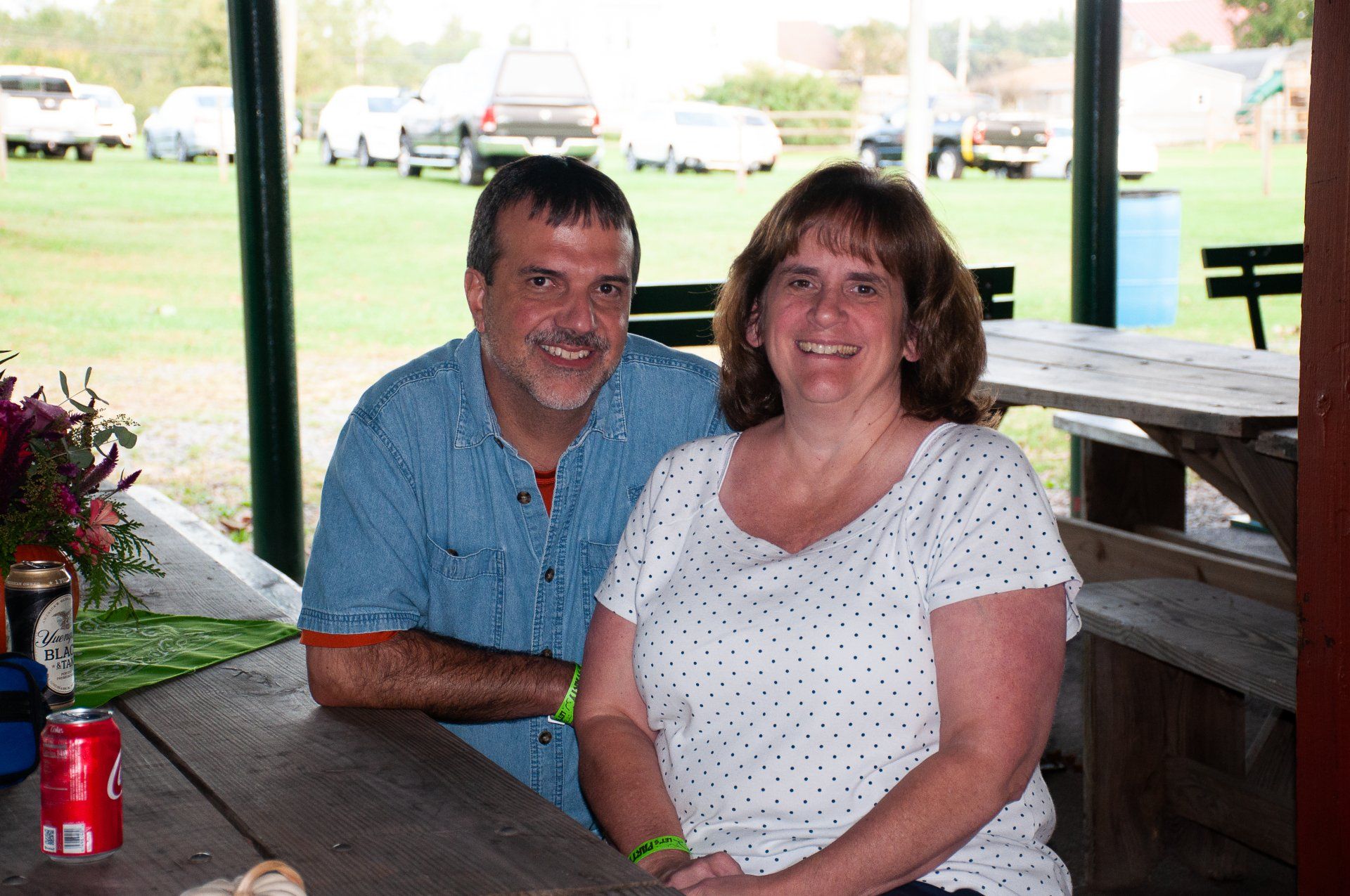 Two people smiling at a picnic table under a pavilion with a grassy field behind them