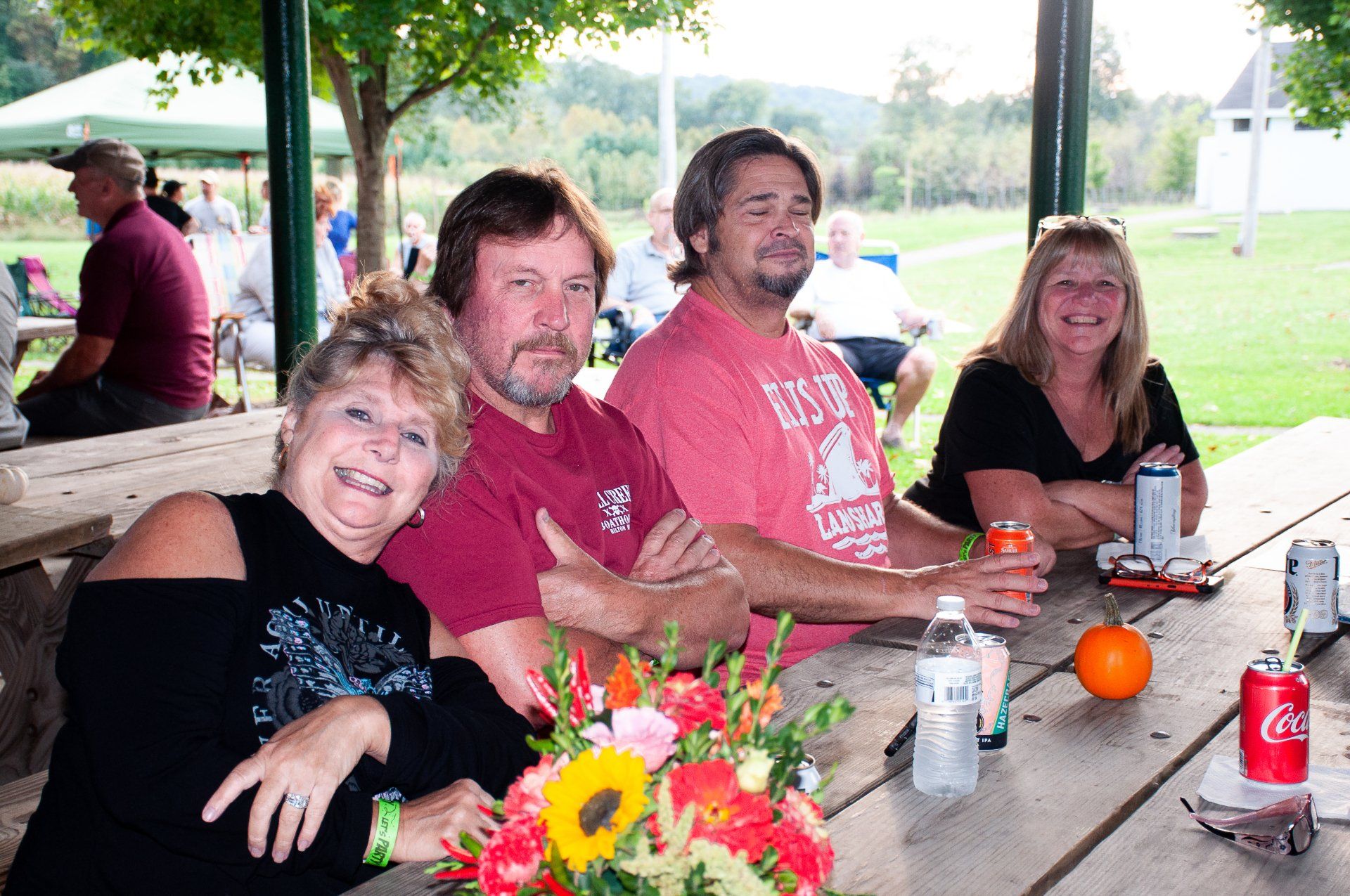 Group of people smiling at a picnic table outdoors with drinks, flowers, and a red soda can