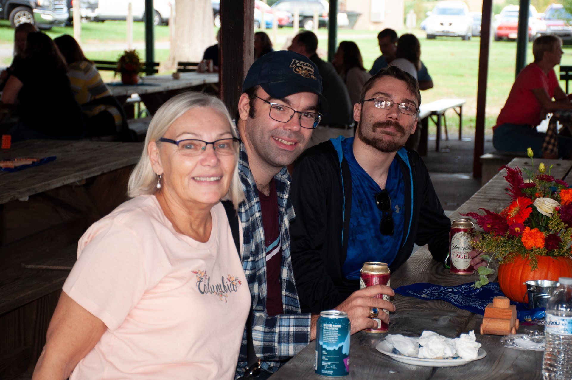 Three people seated at a picnic table in a pavilion, smiling beside drinks and a flower arrangement.