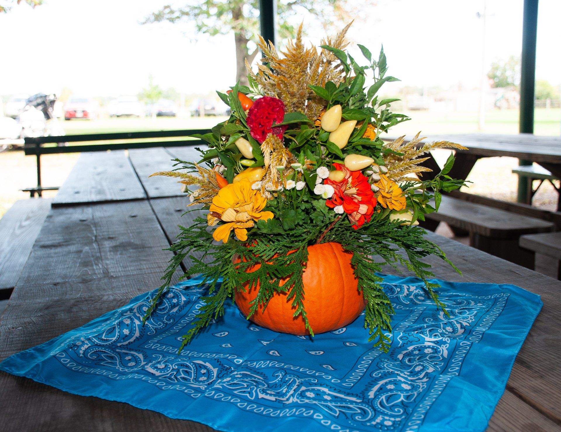 Autumn floral arrangement in a pumpkin vase on a blue bandana-covered picnic table outdoors