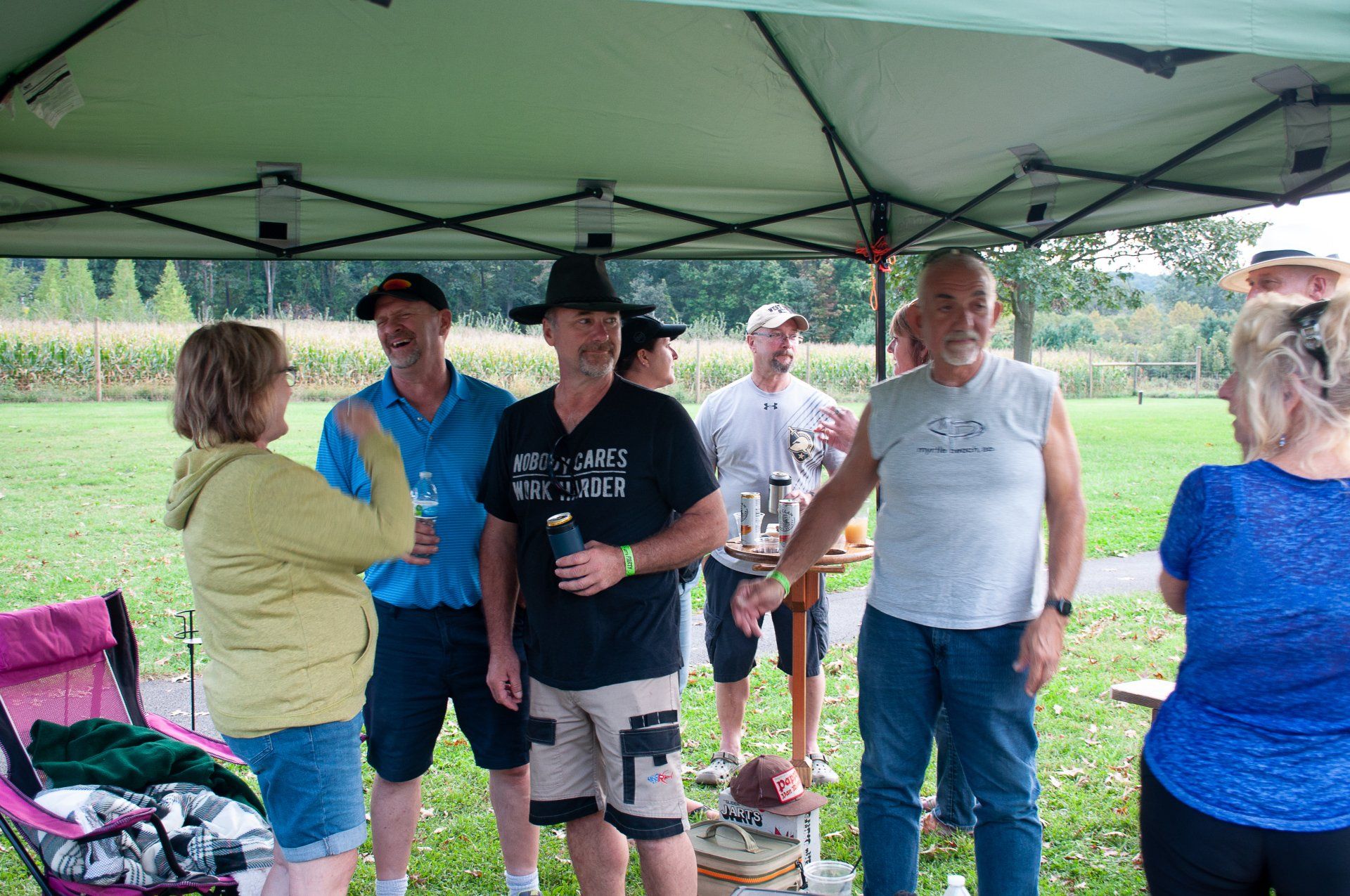 People socializing under a green canopy at an outdoor gathering, holding drinks and chatting.