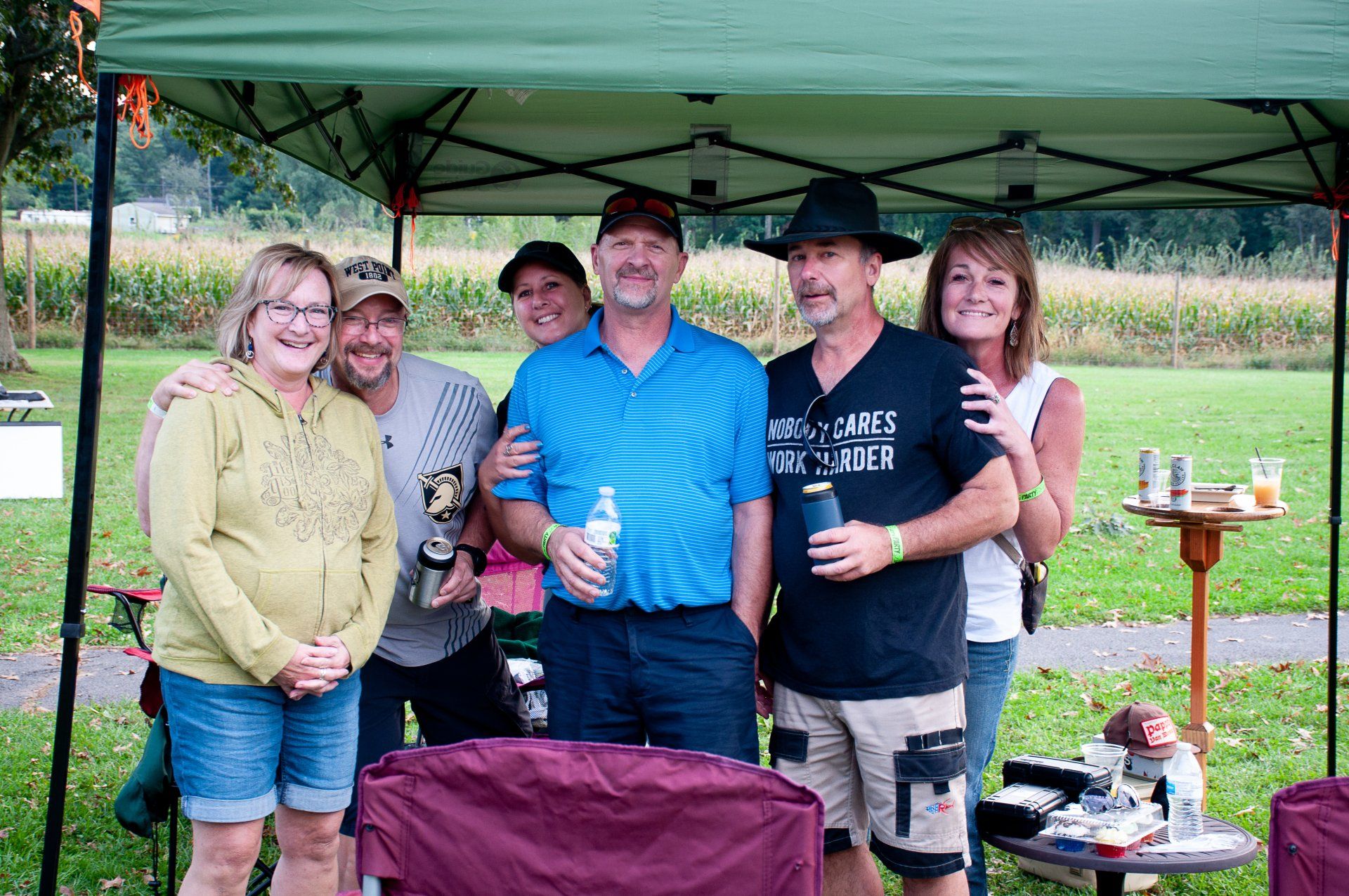 Five people standing under a green canopy at a picnic table, smiling and holding drinks outdoors