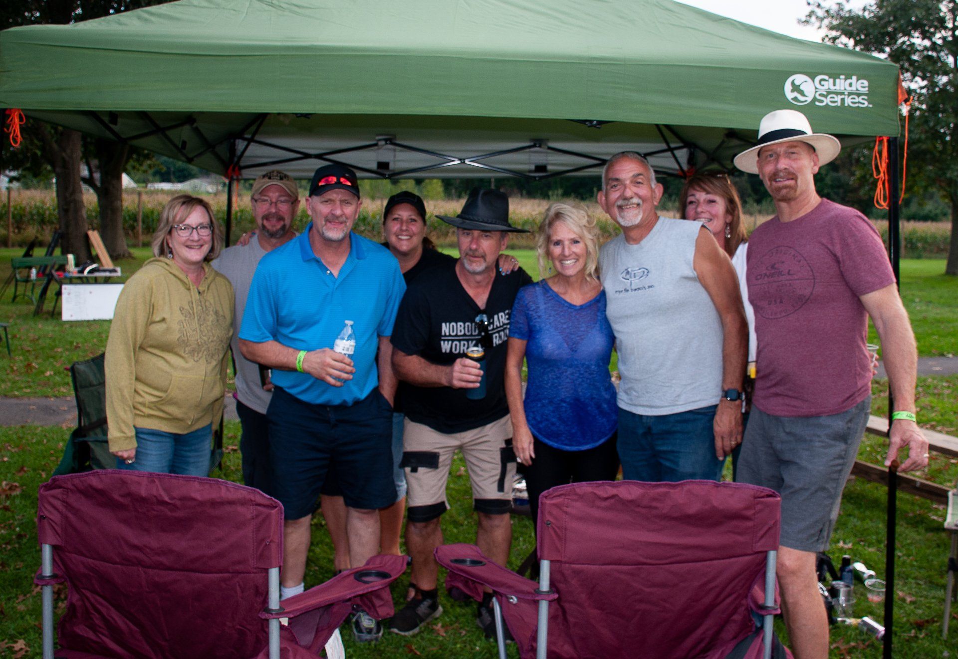 Group of people posing under a green canopy at an outdoor event, smiling and holding drinks.